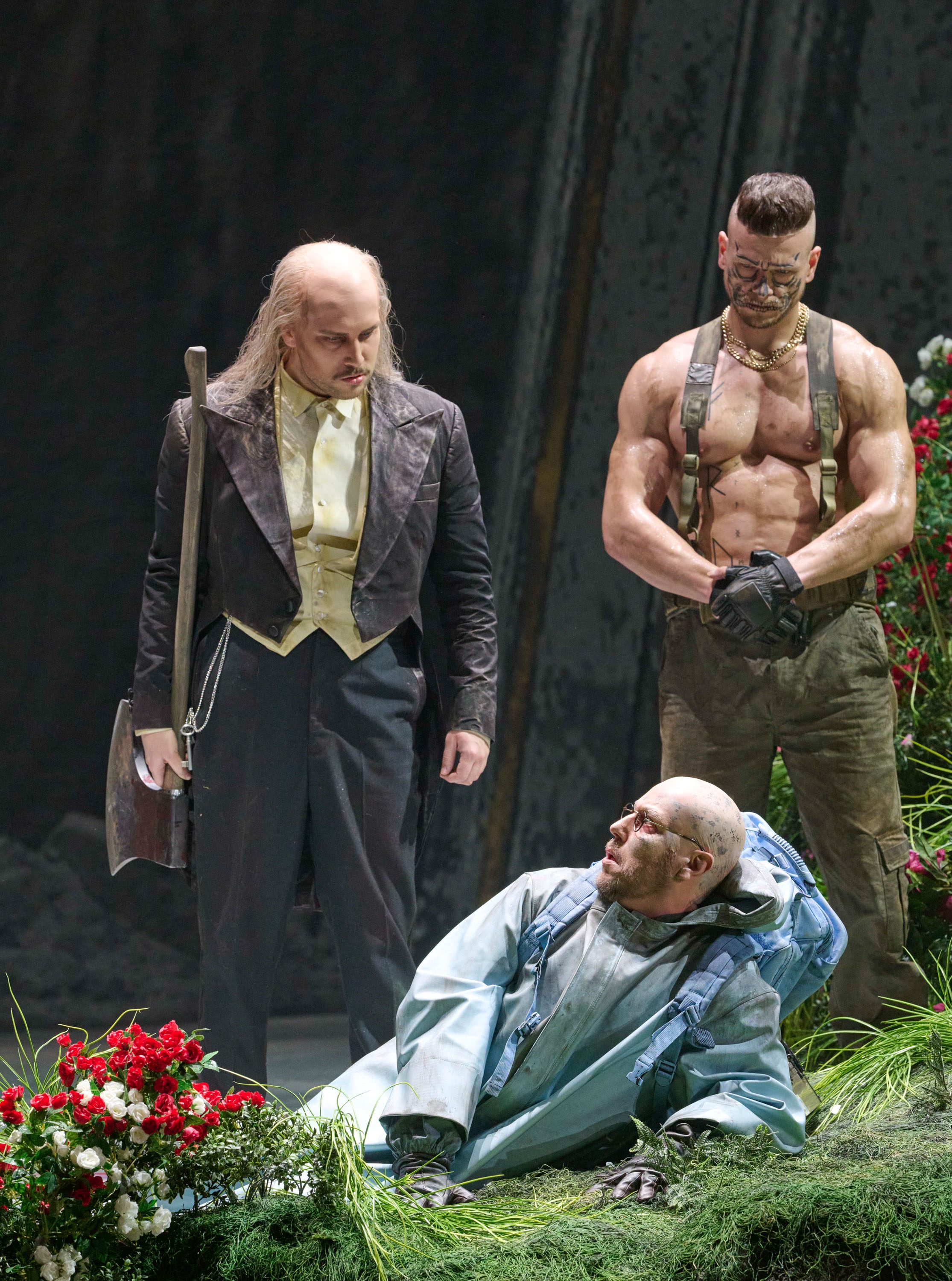 Zwei Männer stehen bedrohlich über einem am Boden sitzenden Mann in blauer Kleidung, umgeben von rotem Blumenschmuck.