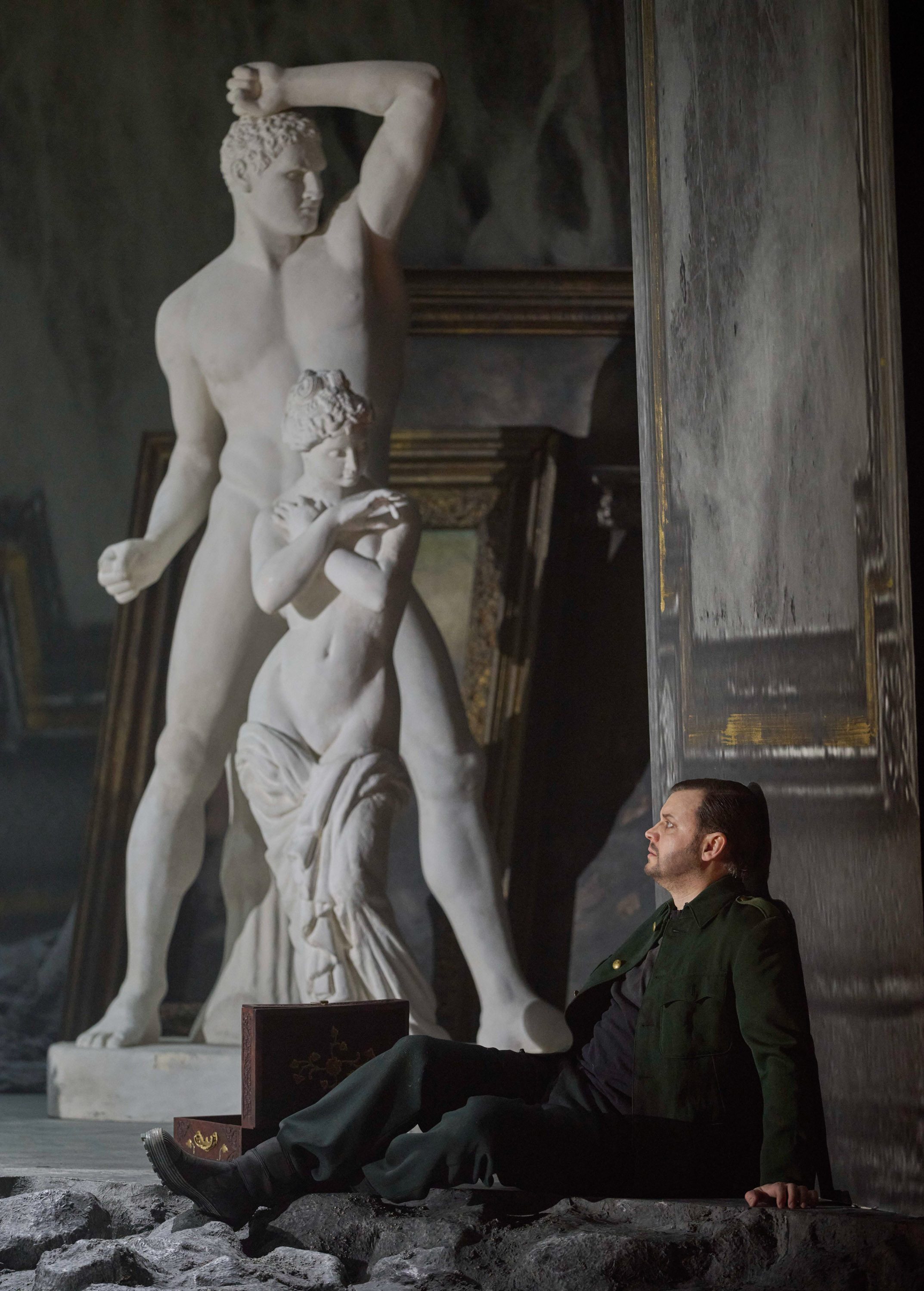 Ein Mann in dunkler Kleidung sitzt nachdenklich vor einer weißen Marmorskulptur und einer geöffneten Truhe auf steinigem Boden.