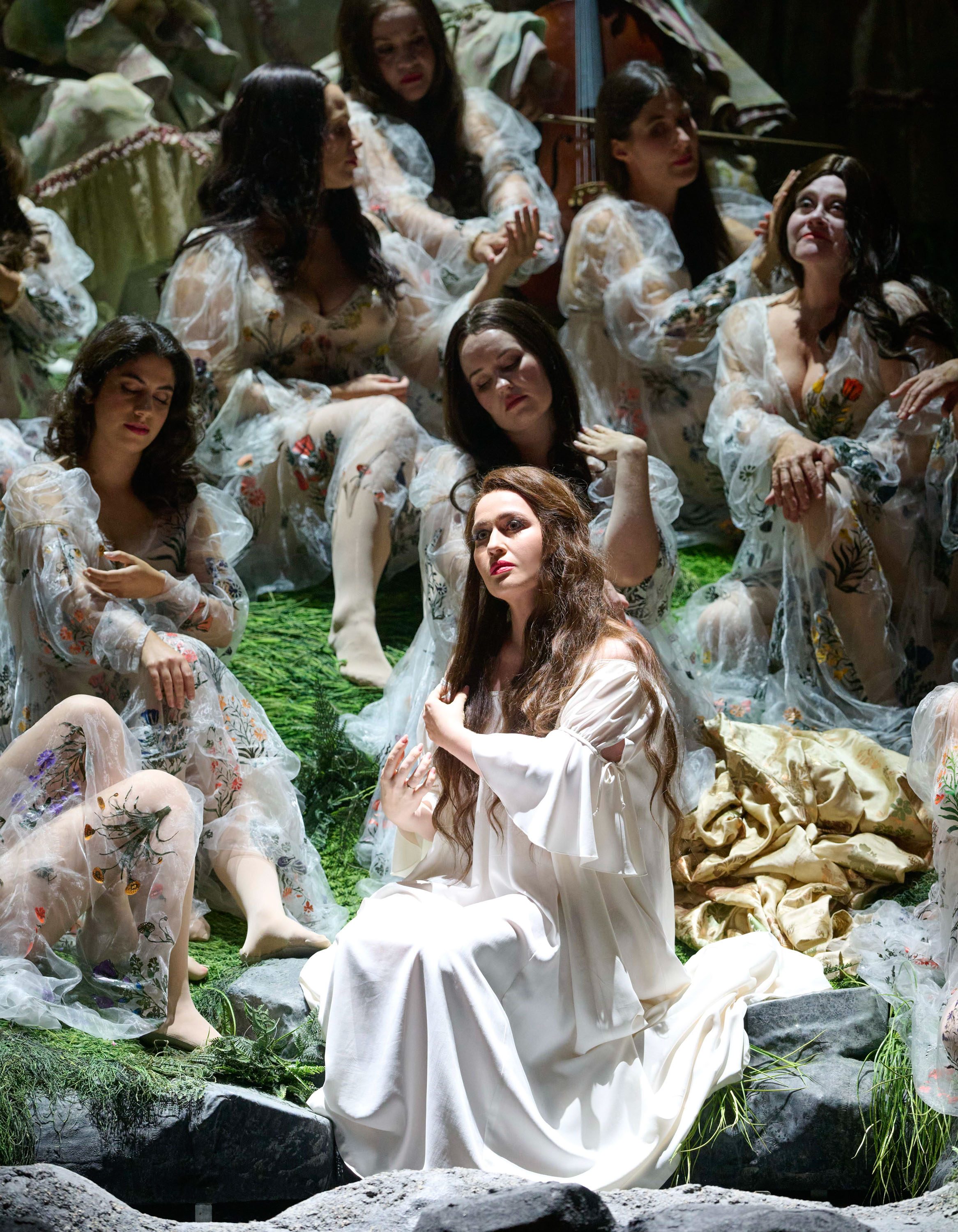 Eine Frau mit langen Haaren sitzt in einem weißen Kleid auf einem grünen Hügel und fährt sich durch die Haare. Um sie herum sitzen weitere Frauen die auch helle Gewänder tragen.