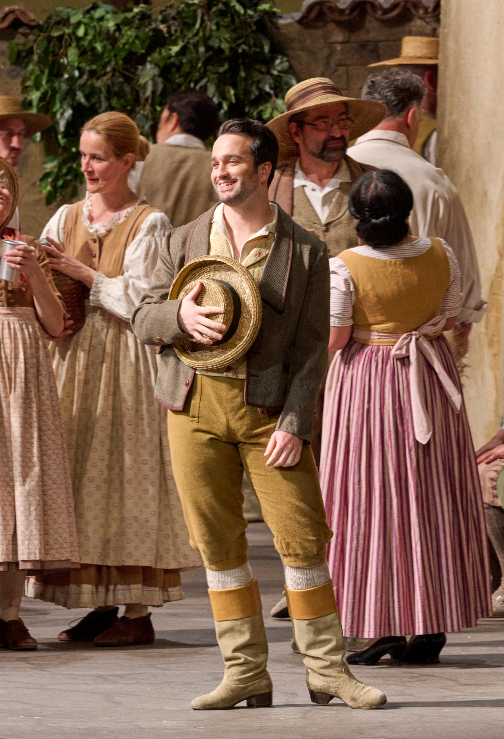 Ein fröhlicher junger Mann mit Strohhut steht lächelnd unter Dorfbewohnern in historischer Kleidung auf einer Bühnenkulisse.
