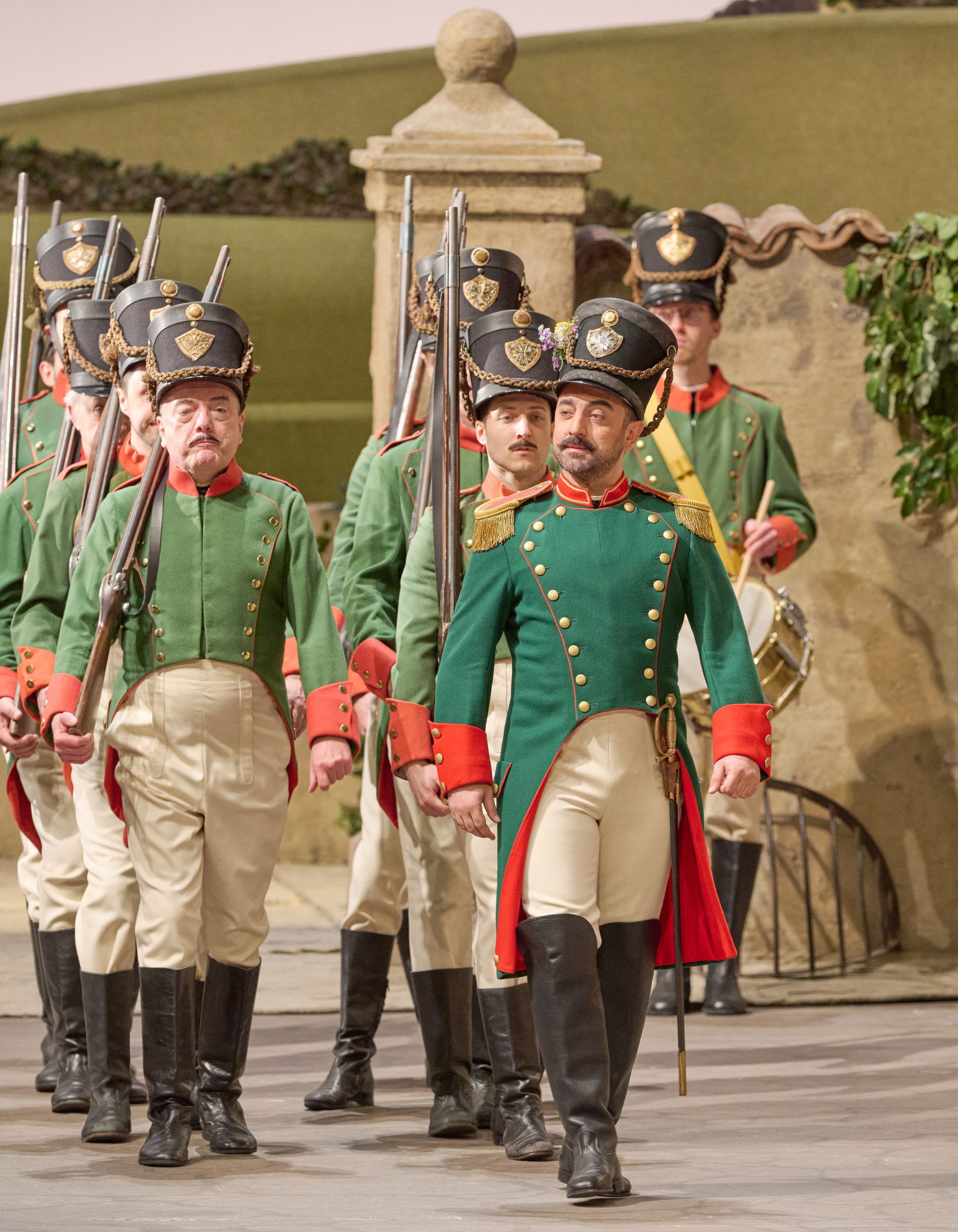 Eine Gruppe von Soldaten in grünen Uniformen marschiert mit Gewehren über eine Opernbühne im historischen Stil.