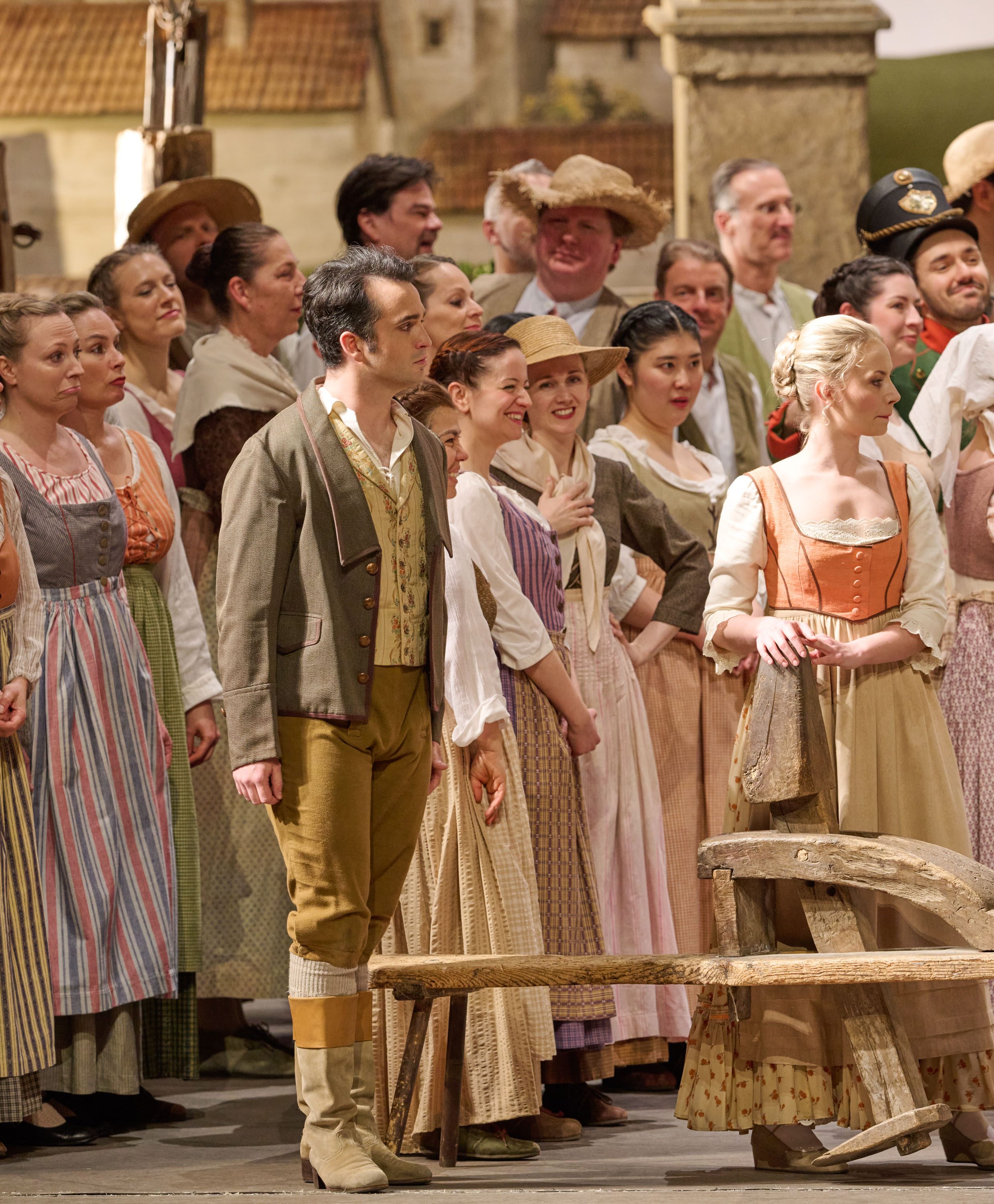 Eine große Gruppe von Dorfbewohnern in bunter historischer Kleidung steht erwartungsvoll auf einer Opernbühne.