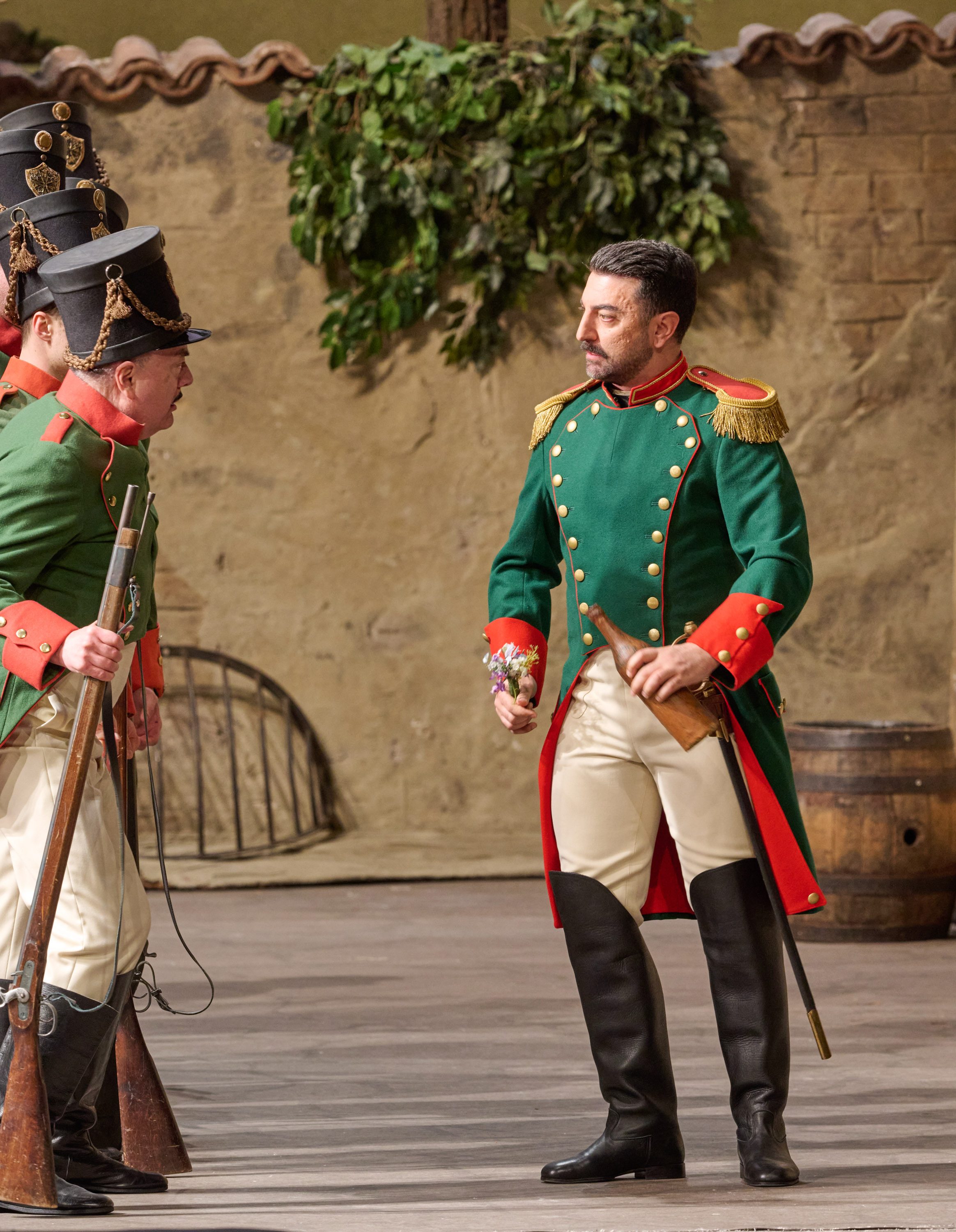 Zwei Soldaten in grüner Uniform unterhalten sich ernsthaft auf einer Bühne mit ländlicher Kulisse.