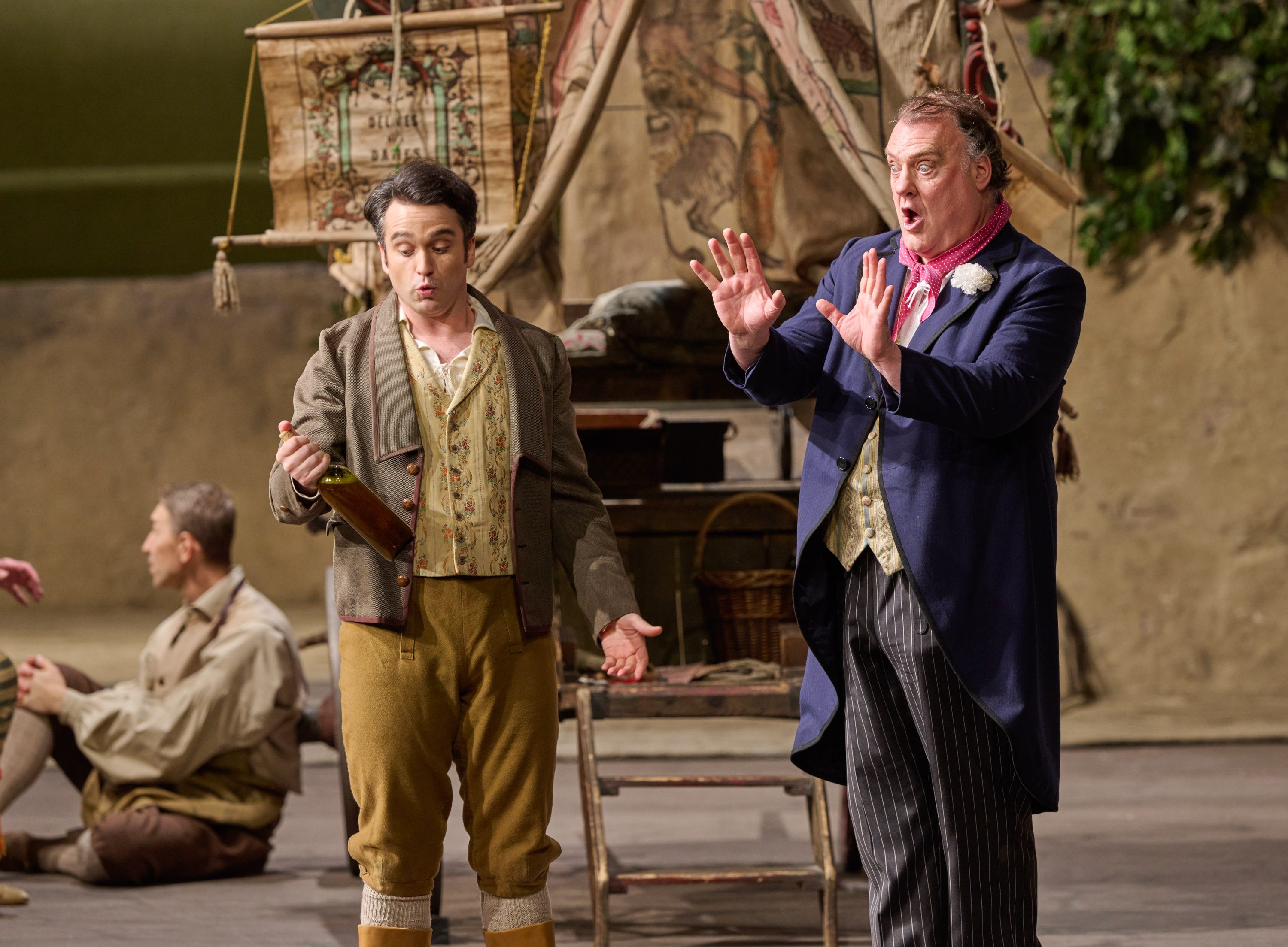 Zwei Männer in unterschiedlicher historischer Kleidung stehen in lebhafter Diskussion auf einer Opernbühne mit Zeltkulisse.