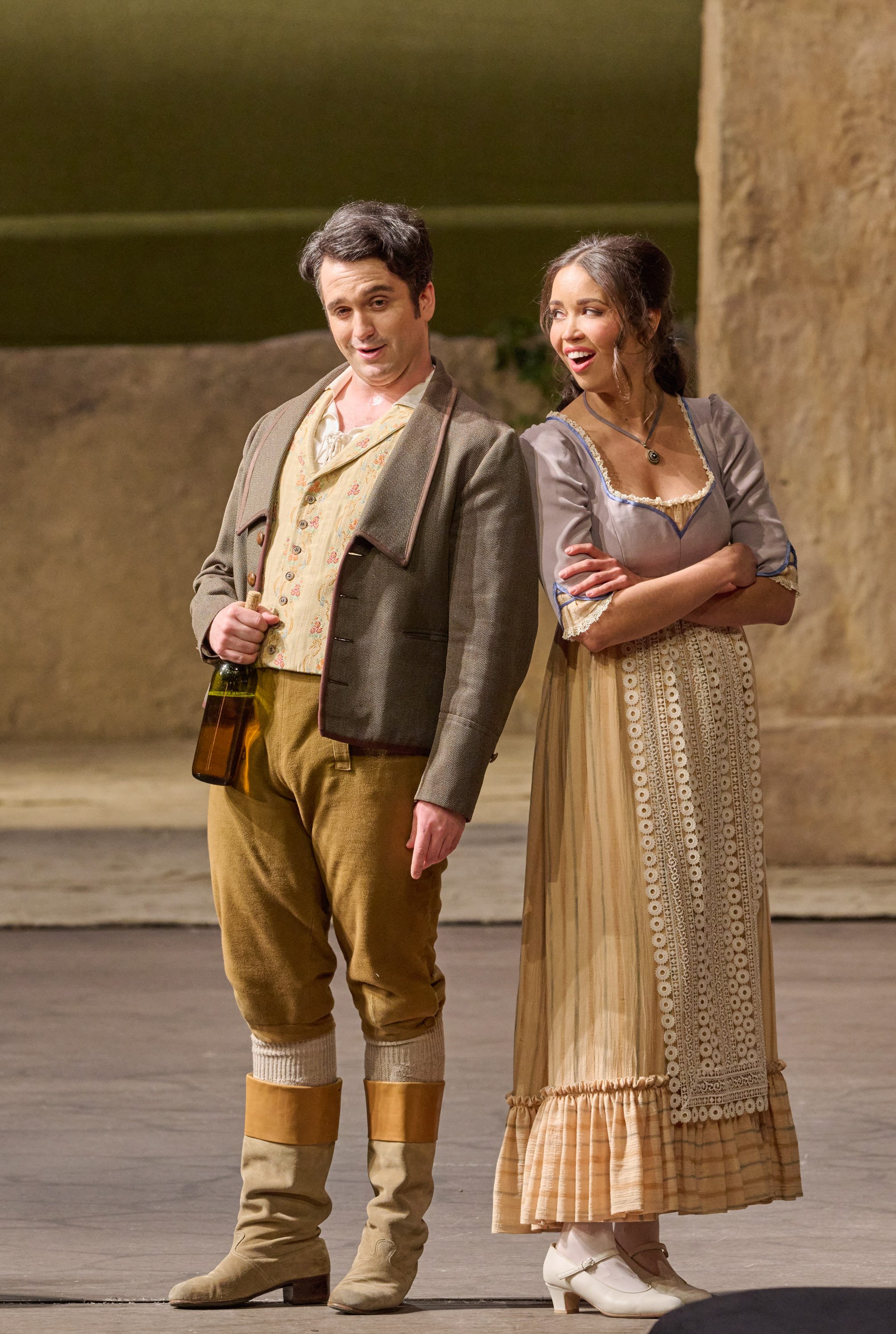 Ein Mann mit Flasche und eine Frau stehen nebeneinander, sie schaut ihn mit verschränkten Armen skeptisch an.