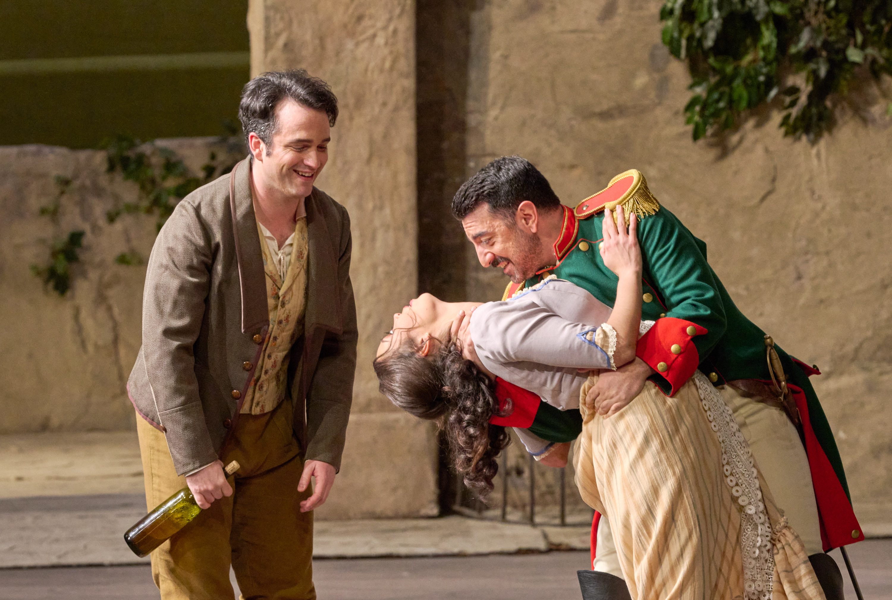 Ein Soldat taucht eine Frau in einer verspielten Tanzpose, während ein lachender Mann mit Flasche danebensteht.