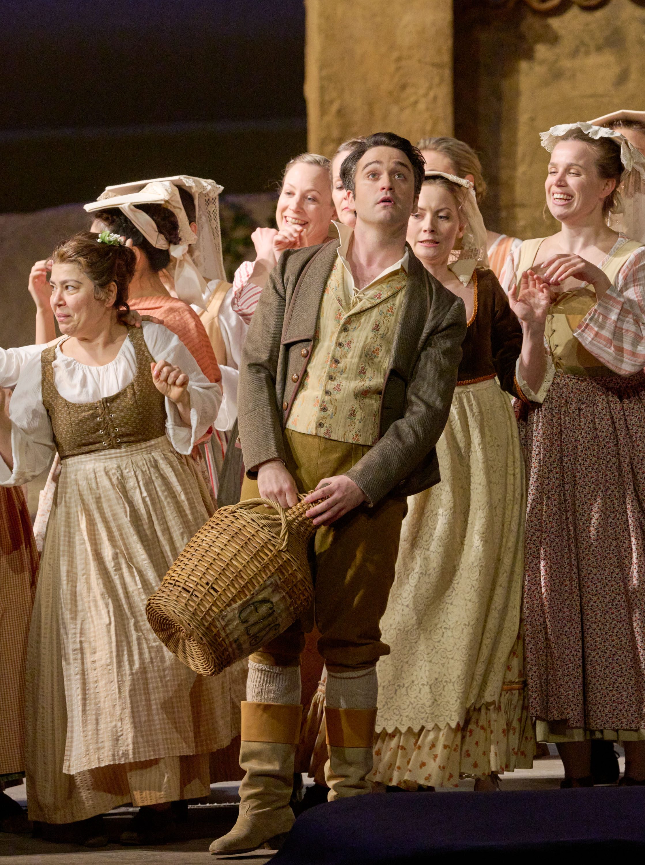 Ein junger Mann mit Korb wird von mehreren Frauen in historischer Kleidung umringt, alle schauen erwartungsvoll nach oben.