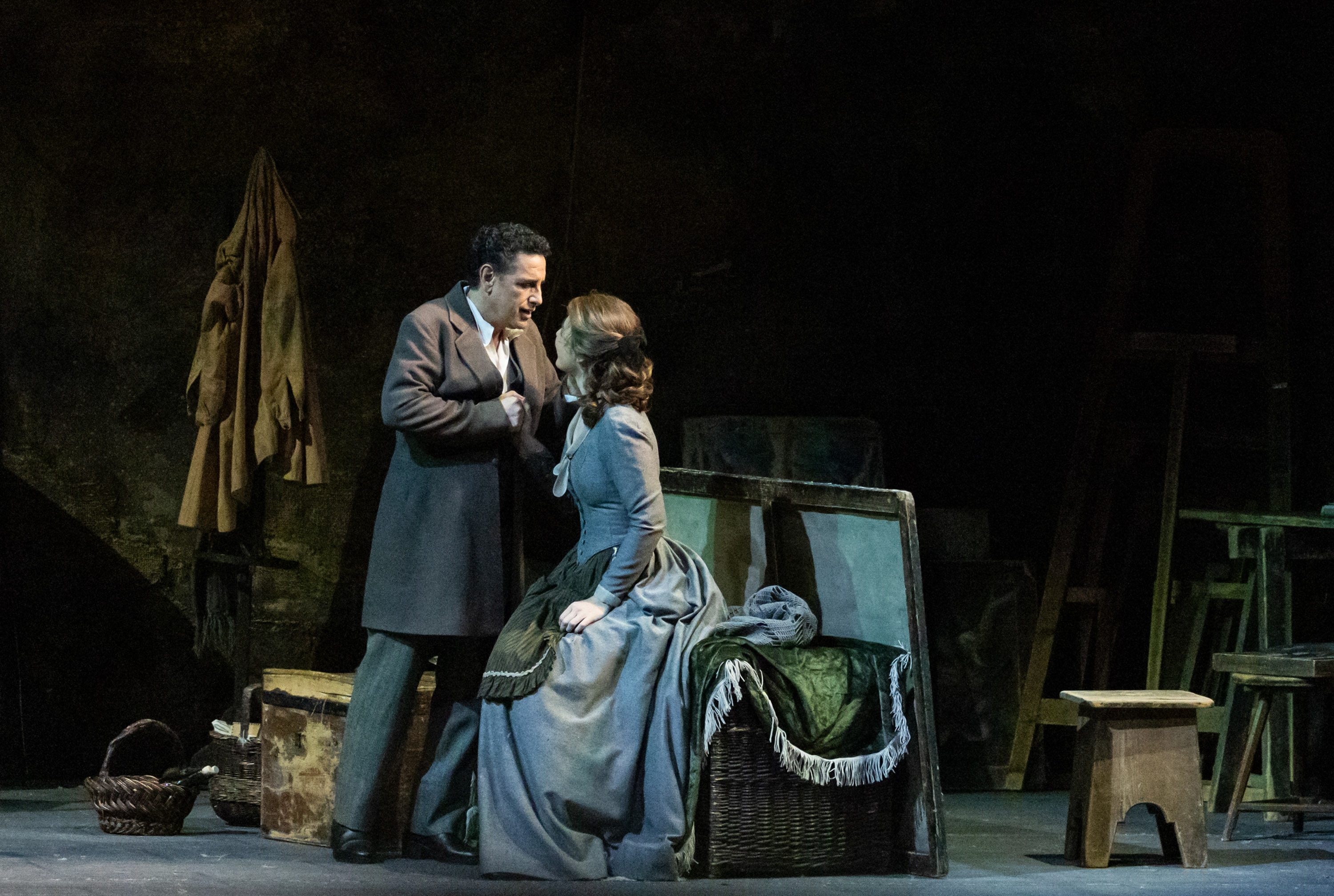 Ein Mann und eine Frau befinden sich in einem dramatischen Austausch. Der Mann steht nahe der Frau, die auf einer Truhe sitzt. Beide tragen historische Kleidung, die auf eine Theateraufführung hinweist. Die Szene zeigt emotionale Intensität in einer dämmerigen Umgebung.