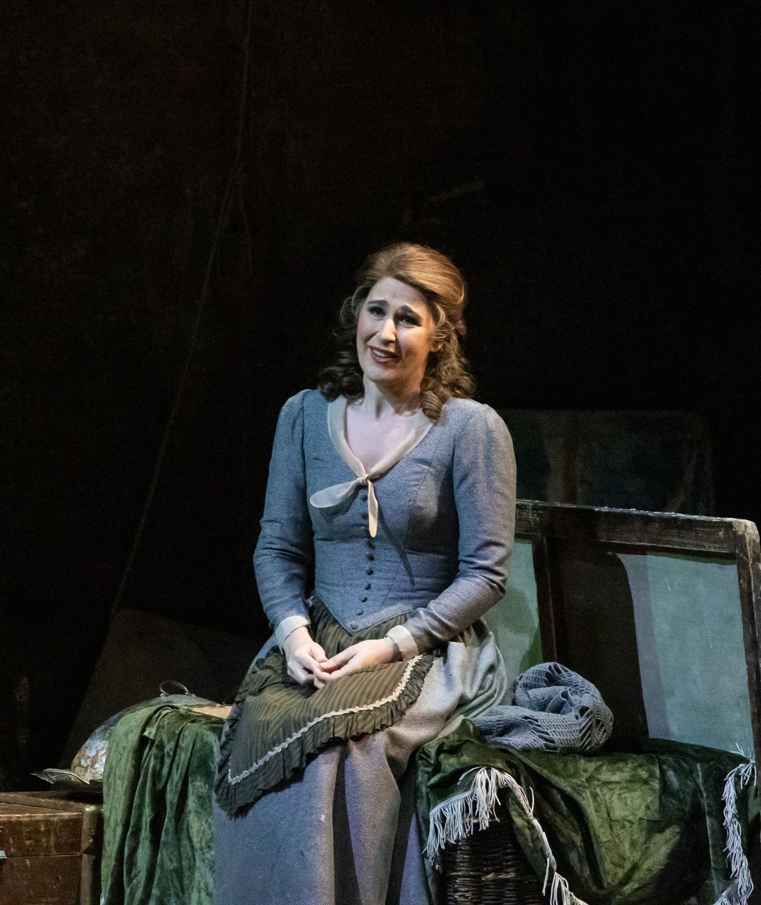 Eine schauspielende Frau sitzt auf einem Koffer, bekleidet mit einem historischen Kleid. Sie hat einen traurigen Ausdruck im Gesicht und schaut emotional. Im Hintergrund sind einige bewegliche Requisiten und einige Textilien sichtbar. Die Szene vermittelt einen Ausdruck von Melancholie.