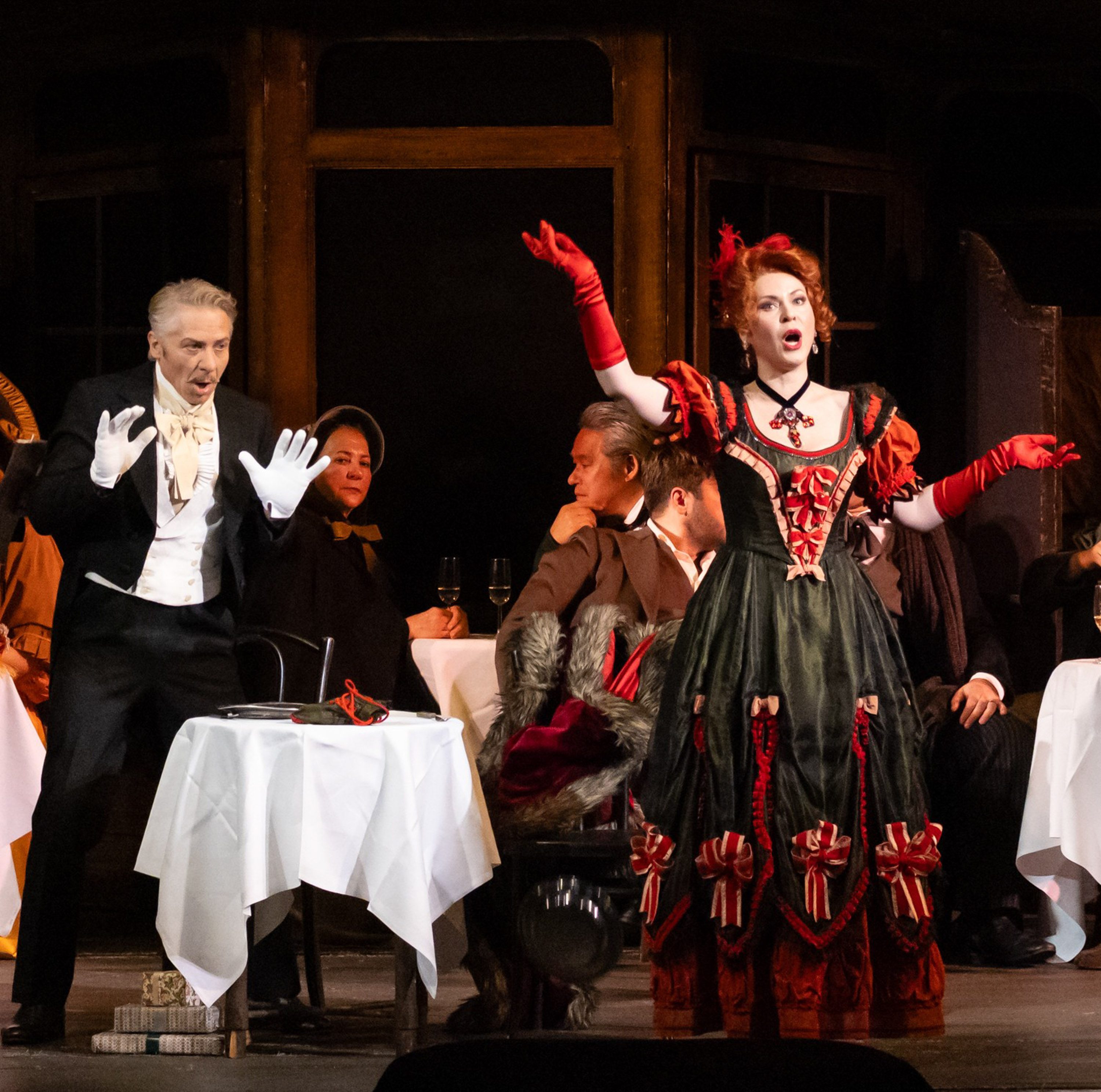 Eine Frau in einem prunkvollen, roten Kleid mit schwarzen Akzenten steht auf einer Bühne und singt leidenschaftlich. Neben ihr steht ein Mann in smoking, der überrascht reagiert. Im Hintergrund sitzen weitere Personen an Tischen und beobachten die Szene.