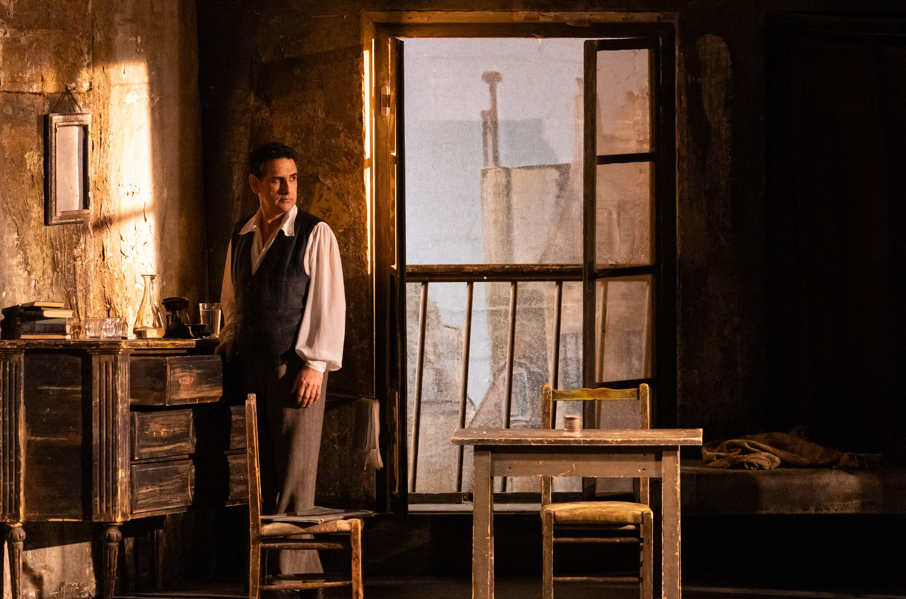 Der Bildinhalt zeigt einen Mann in eleganter Kleidung, der in einem heruntergekommenen Raum mit einer offenen Fenster steht. Auf einem Holzschreibtisch sind ein Glas und einige Bücher zu sehen. Das Licht fällt sanft durch das Fenster und schafft eine nachdenkliche Atmosphäre.