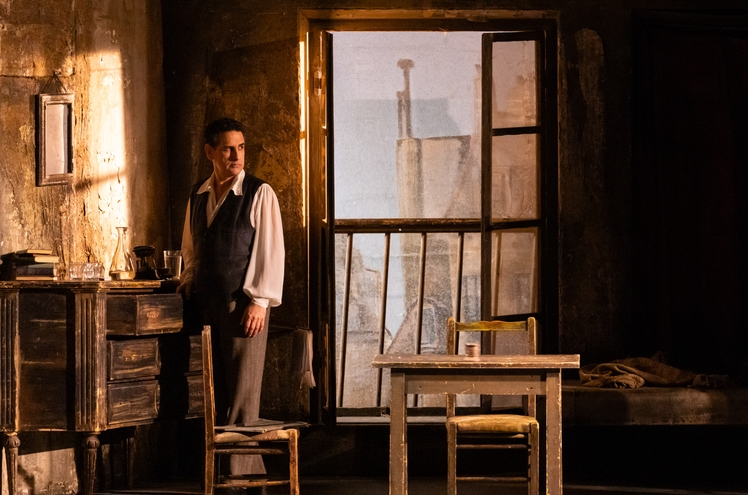 A man in historical attire stands near a wooden dresser in a dimly lit room. Sunlight filters through an open window, illuminating his thoughtful expression. A table with a small cylindrical object is nearby, and a chair is positioned at the table.
