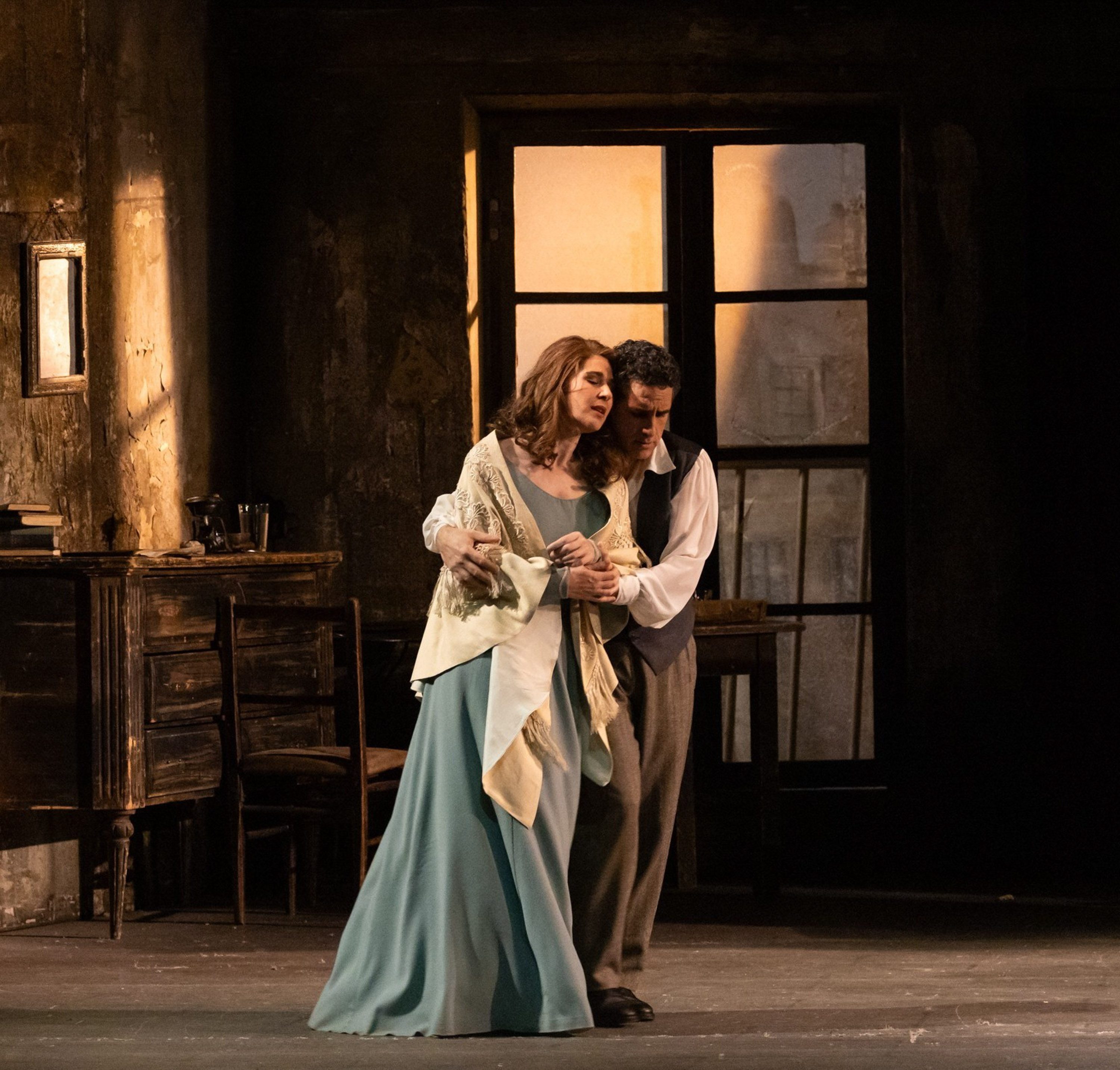 Ein Paar steht eng umschlungen auf einer Theaterbühne. Die Frau trägt ein elegantes, fließendes Kleid und eine Stola. Beide zeigen Emotionen, die Situation wirkt dramatisch. Im Hintergrund sind dunkle Wände und ein Fenster mit schwachem Licht sichtbar.