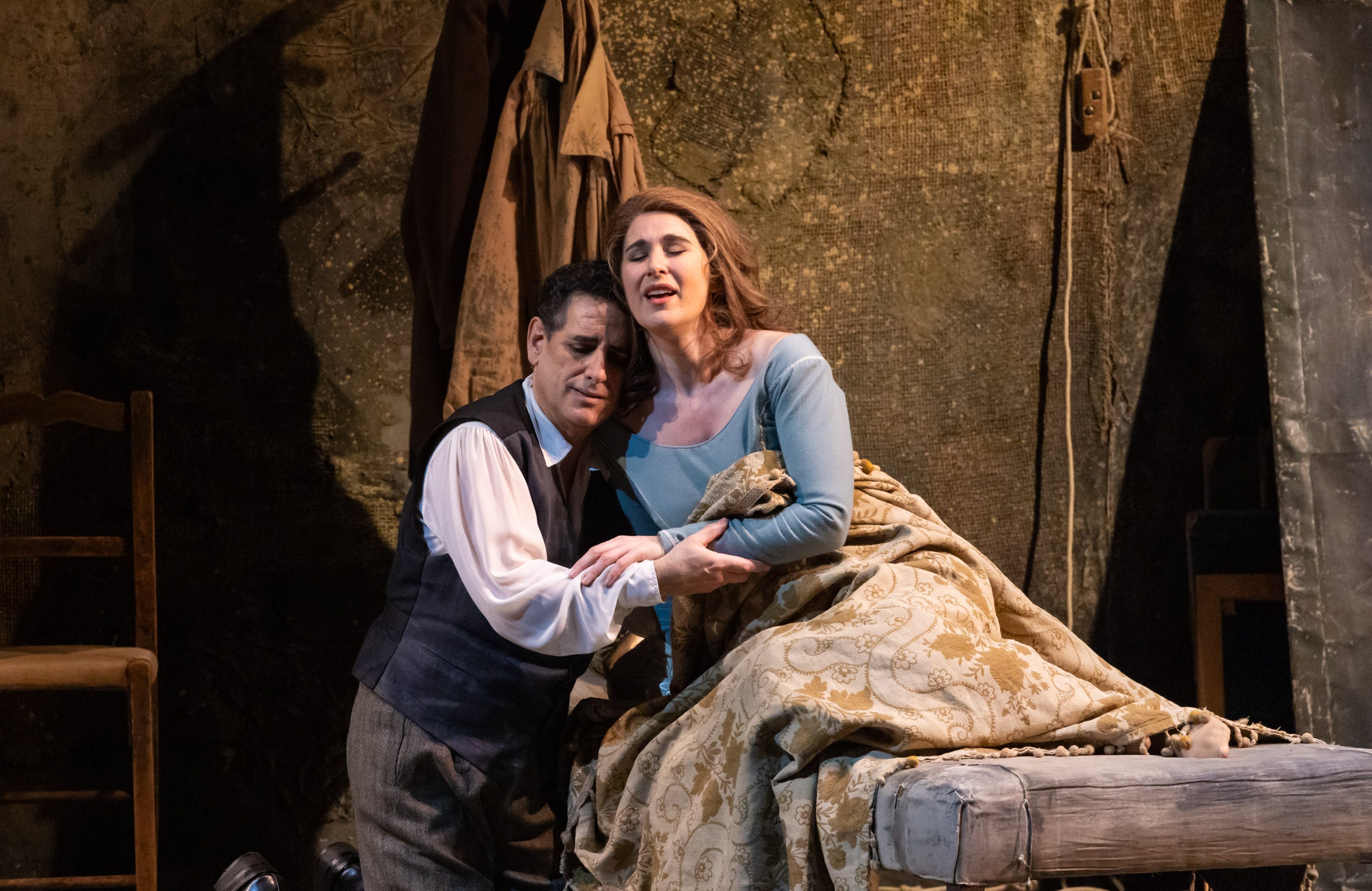 Zwei Darsteller in einem dramatischen Moment auf einer Bühne. Der männliche Charakter umarmt die weibliche Figur, die auf einem Bett sitzt, und beide zeigen starke Emotionen. Der Hintergrund ist schlicht und unauffällig, was die Intensität ihrer Darstellung unterstreicht.