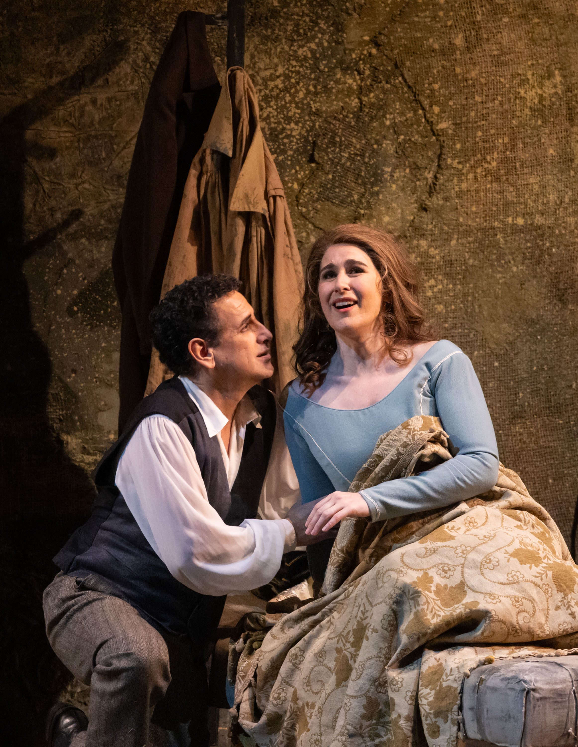 Ein Mann und eine Frau sitzen in einem stimmungsvollen Raum. Der Mann blickt intensiv auf die Frau, während sie ihn anlächelt. Sie trägt ein elegantes, hellblaues Kleid und ist mit einer Decke umhüllt. Die Szene vermittelt eine emotionale Verbindung zwischen den beiden.