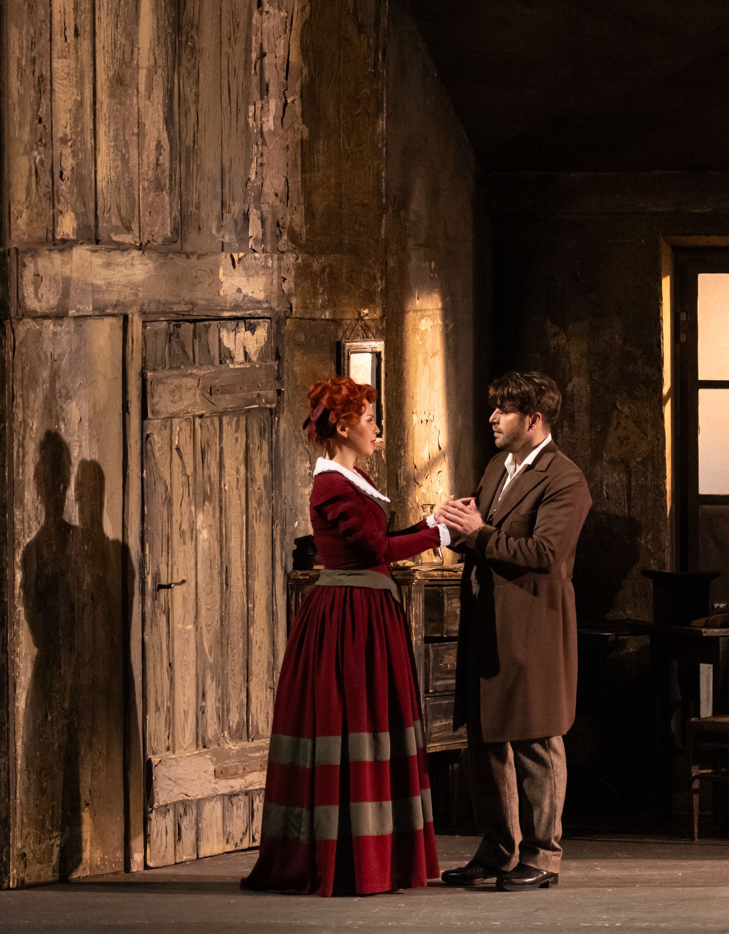 Das Bild zeigt ein Schauspielszene mit einem Mann und einer Frau, die sich gegenüberstehen und Händchen halten. Die Frau trägt ein rot gestreiftes Kleid, während der Mann einen langen Mantel trägt. Die Szene ist von einer rustikalen Kulisse mit Holzvertäfelungen umgeben.