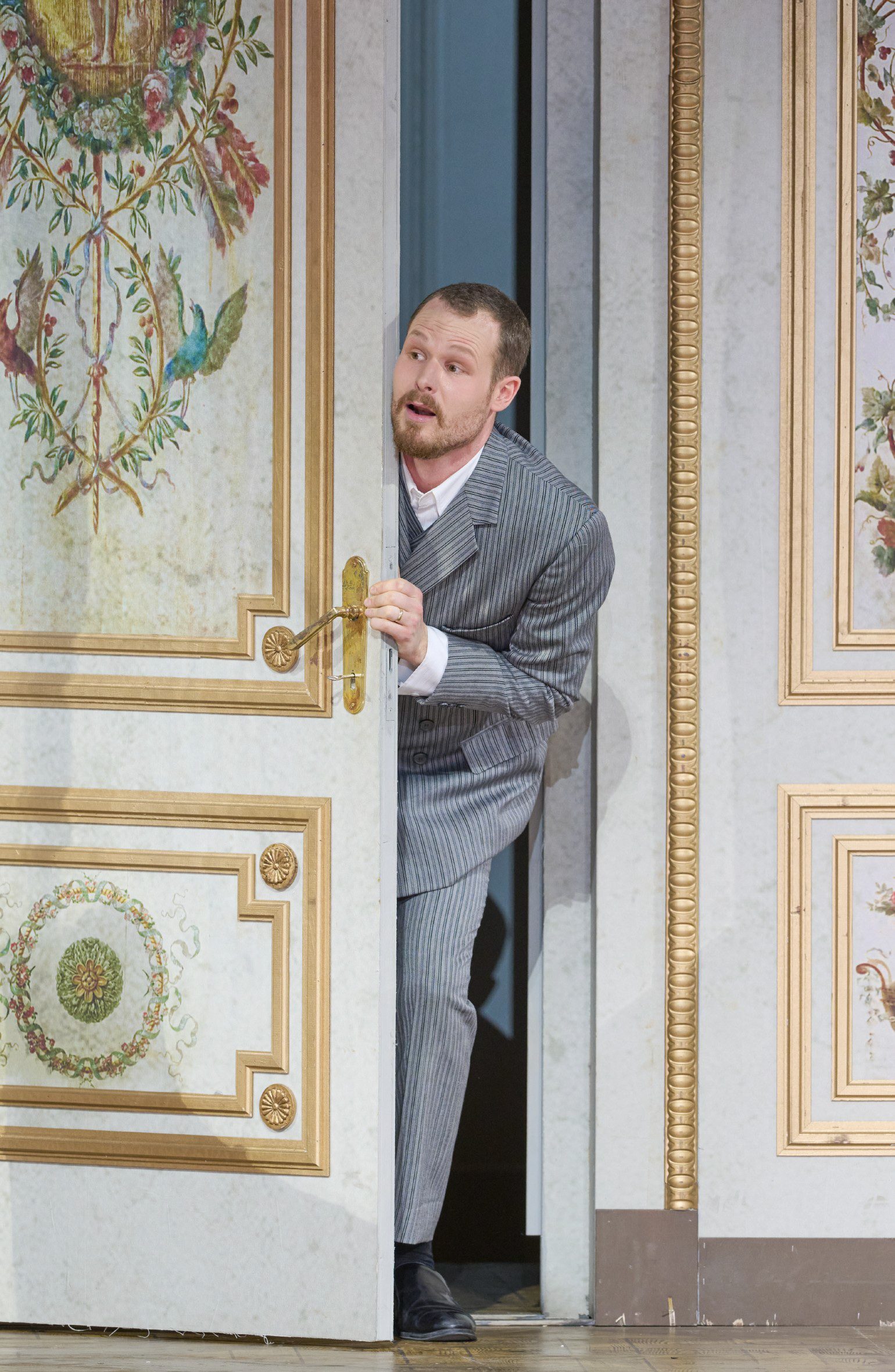 Ein Mann im grauen Anzug lugt neugierig durch eine halb geöffnete, prunkvoll verzierte Tür in einem klassisch dekorierten Raum.