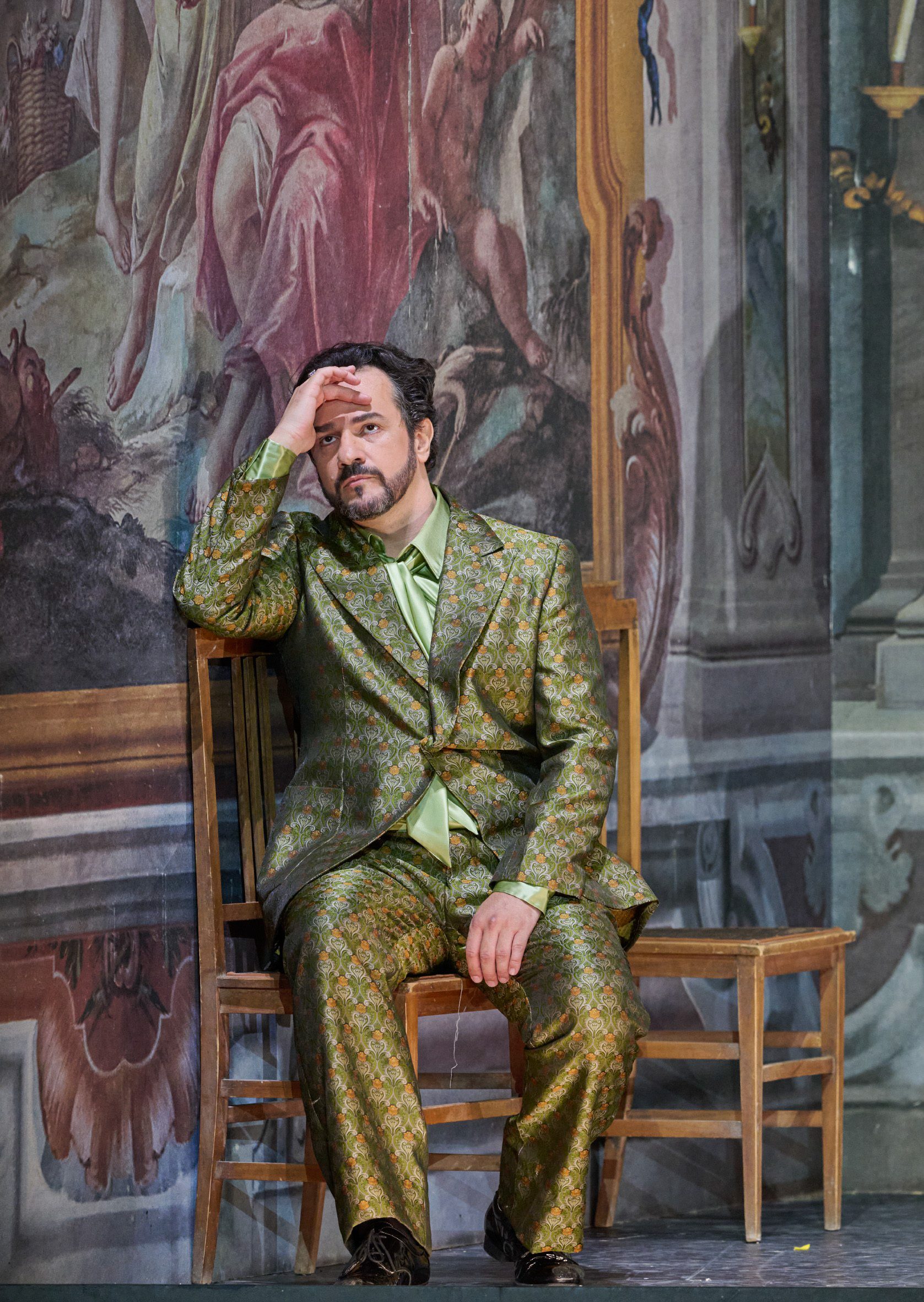 Ein Mann in glänzend grün-gemustertem Anzug sitzt nachdenklich auf einem Stuhl vor einer großflächigen barocken Wandmalerei.