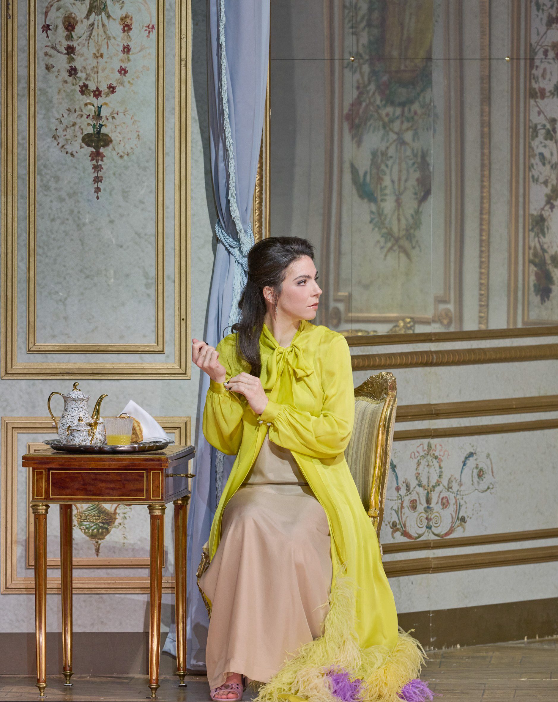 Eine Frau im eleganten gelben Mantel sitzt in einem reich dekorierten Raum auf einem Sessel und schaut gespannt zur Seite, während auf dem Tisch neben ihr ein Teeservice steht.