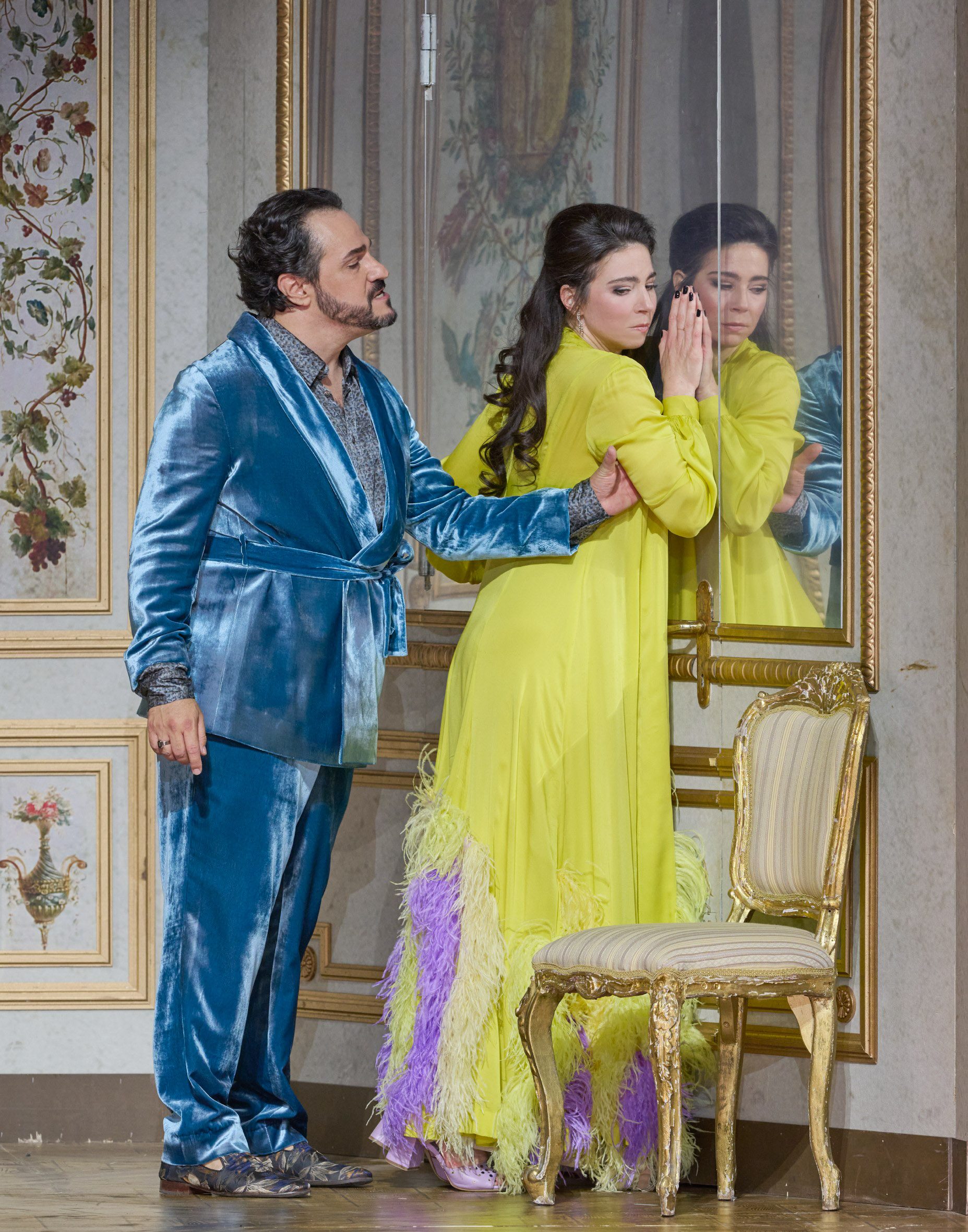 Ein Mann im blauen Samtanzug spricht eindringlich mit einer Frau im gelben Kleid, die sich mit geschlossenen Augen und gesenktem Kopf gegen eine spiegelnde Wand lehnt.