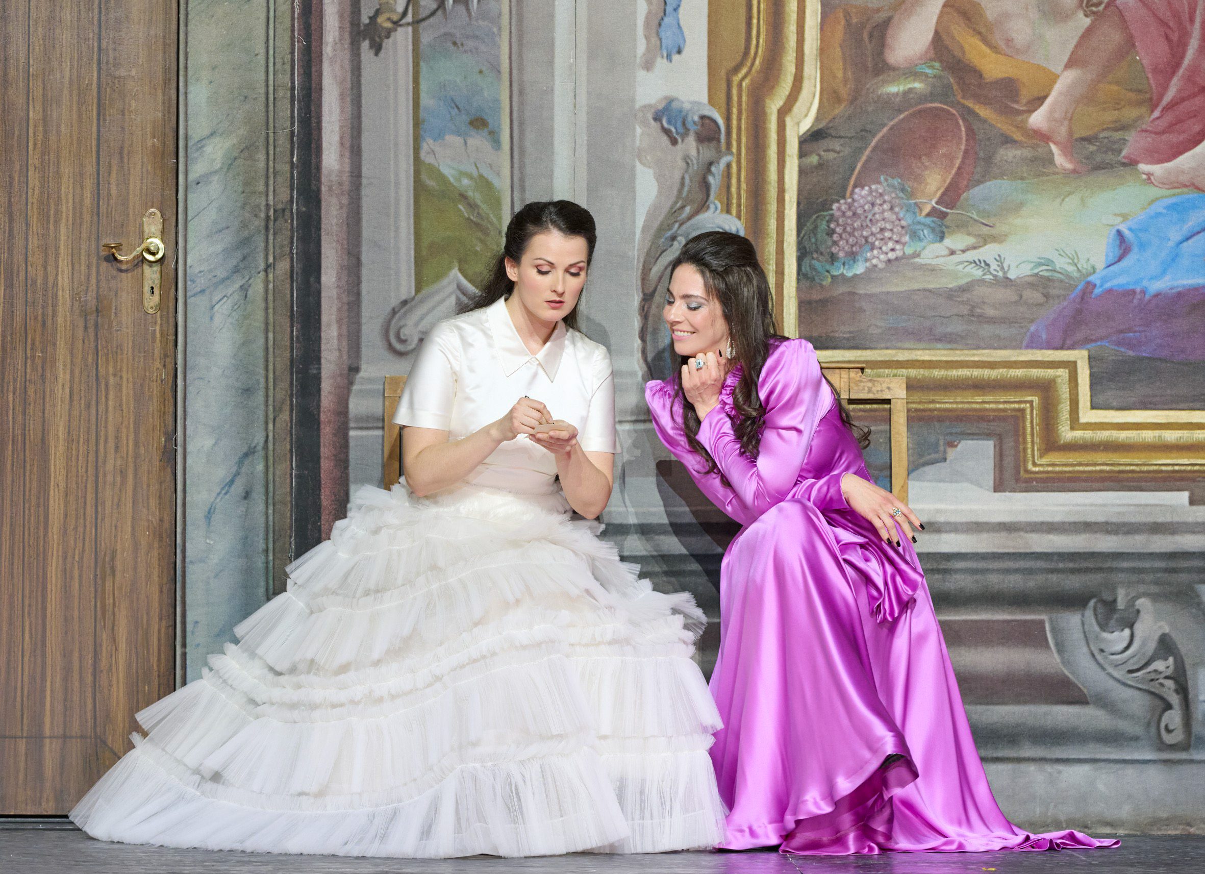 Zwei Frauen sitzen nebeneinander vor einer barocken Wandmalerei; die eine trägt ein weißes Rüschenkleid und zeigt der anderen in einem glänzenden pinken Kleid etwas in ihren Händen.