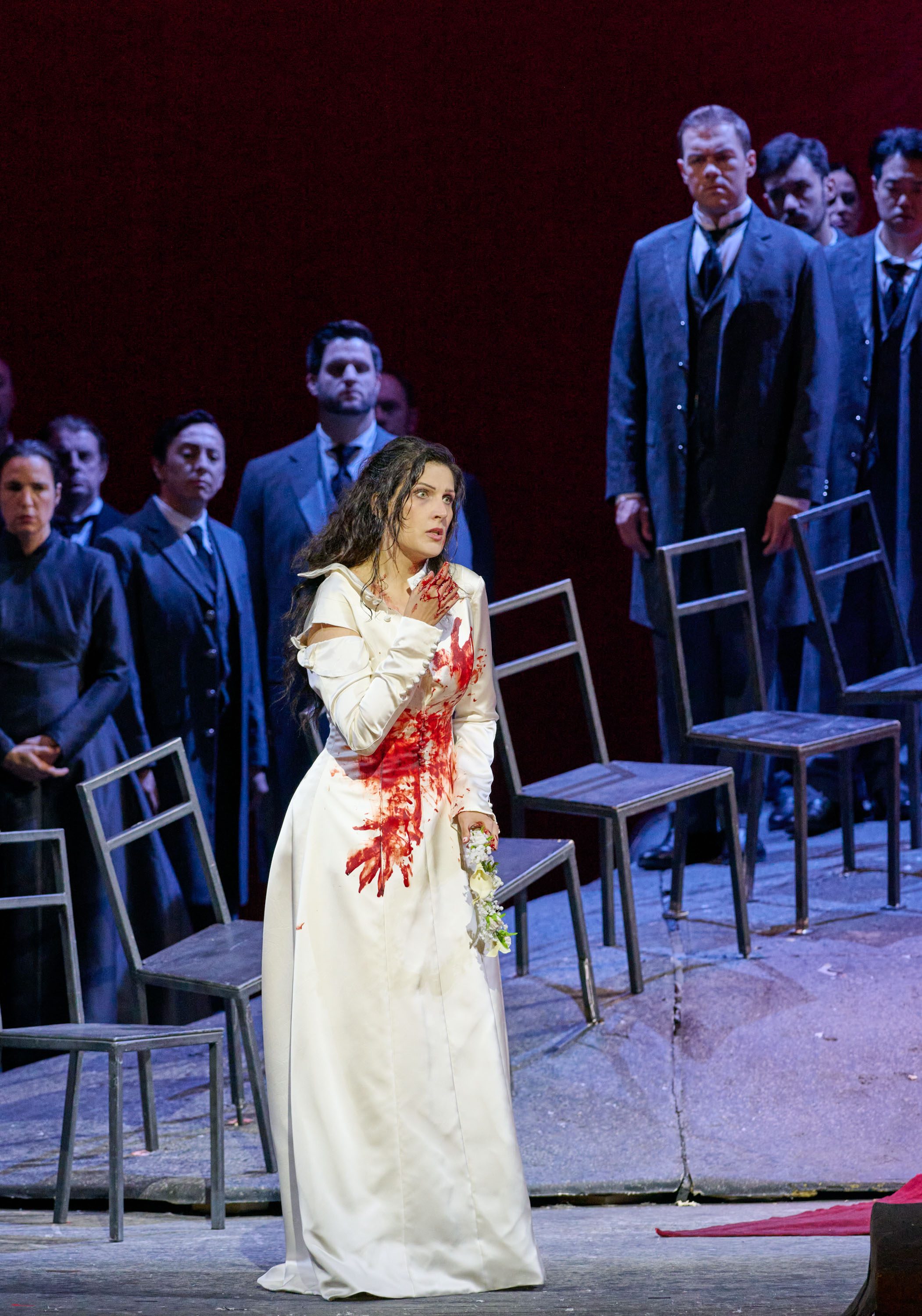 Eine Frau in einem blutverschmierten weißen Kleid steht verzweifelt auf einer Bühne. Sie greift sich an die Brust und hält einen Strauß Blumen in der Hand. Im Hintergrund stehen mehrere Personen in dunkler Kleidung und beobachten sie.
