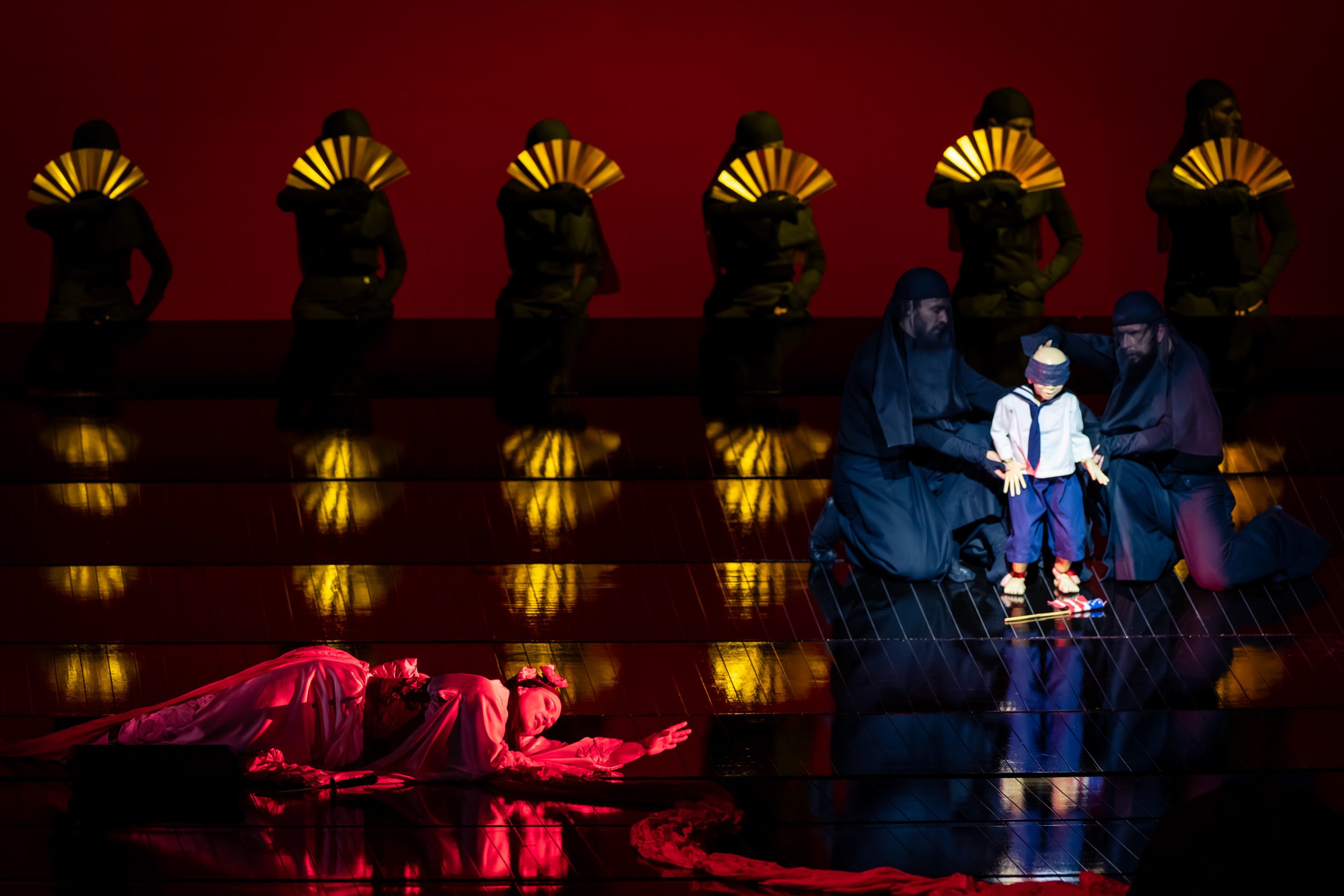 A dramatic theater scene features a child-like puppet standing on stage, surrounded by two figures in dark attire, while a figure in white lies on the floor. Background characters, holding fans, are partially obscured by a vibrant red setting, creating an intense atmosphere.