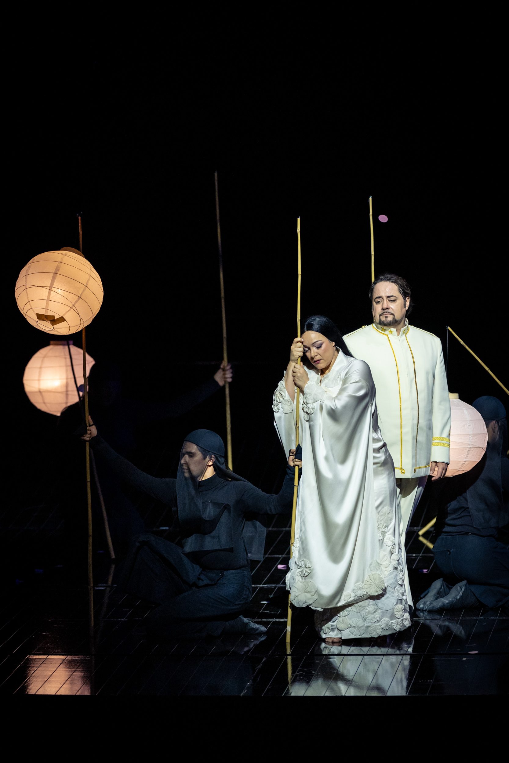 A scene from a dramatic performance features two central characters: a dressed in a flowing white gown with intricate details, and a man in a white suit with gold accents. They are surrounded by performers in dark attire, holding bamboo sticks and illuminated lanterns, creating a moody atmosphere.