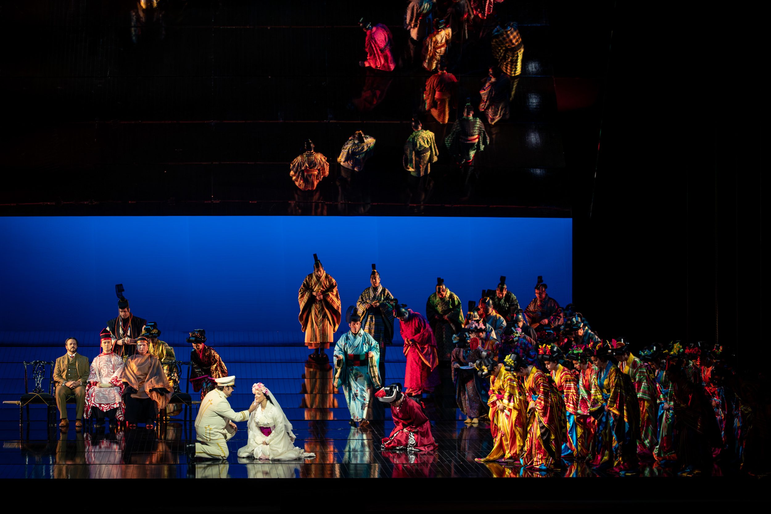 A theatrical performance is depicted, featuring a central couple in traditional attire, with the male character kneeling and interacting with the female character. Surrounding them are numerous performers in vibrant costumes, set against a blue backdrop, creating a dynamic and colorful atmosphere.