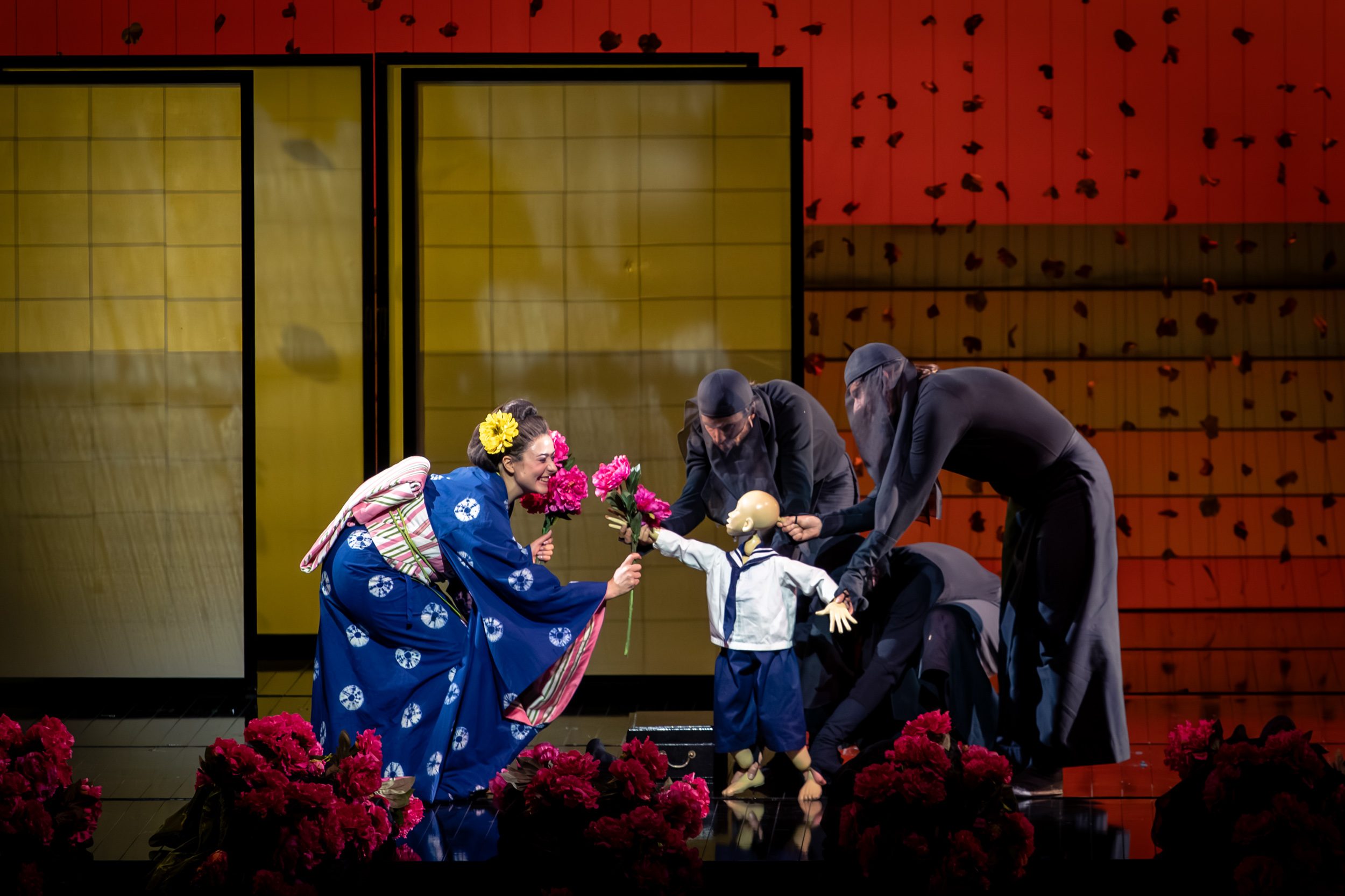 A performer in a blue kimono hands flowers to a doll dressed in a sailor suit. Four figures in dark costumes assist and surround the doll. The stage features a colorful backdrop and floral decorations, creating a vibrant and theatrical scene.