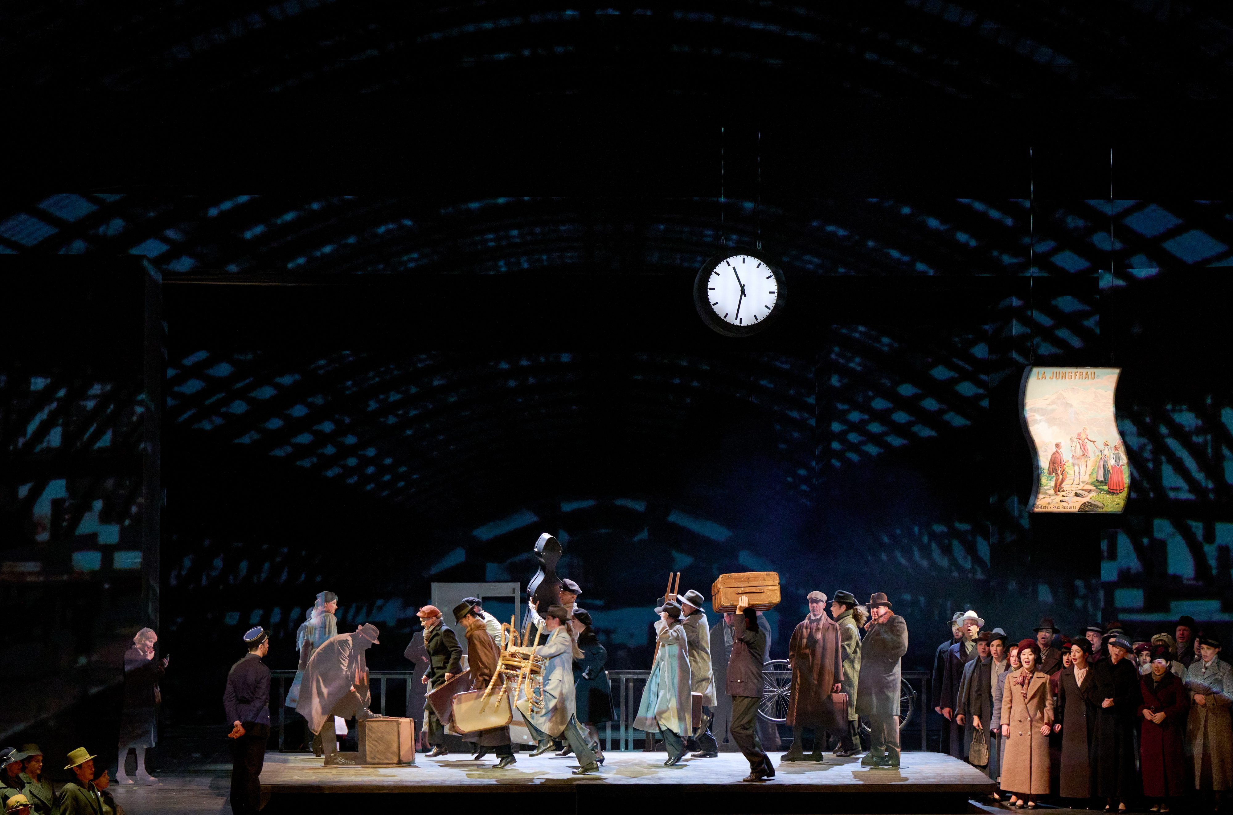 Eine Gruppe von Menschen in historischen Kostümen steht mit Gepäck unter einer großen Bahnhofsuhr auf einer Bühnenkulisse.