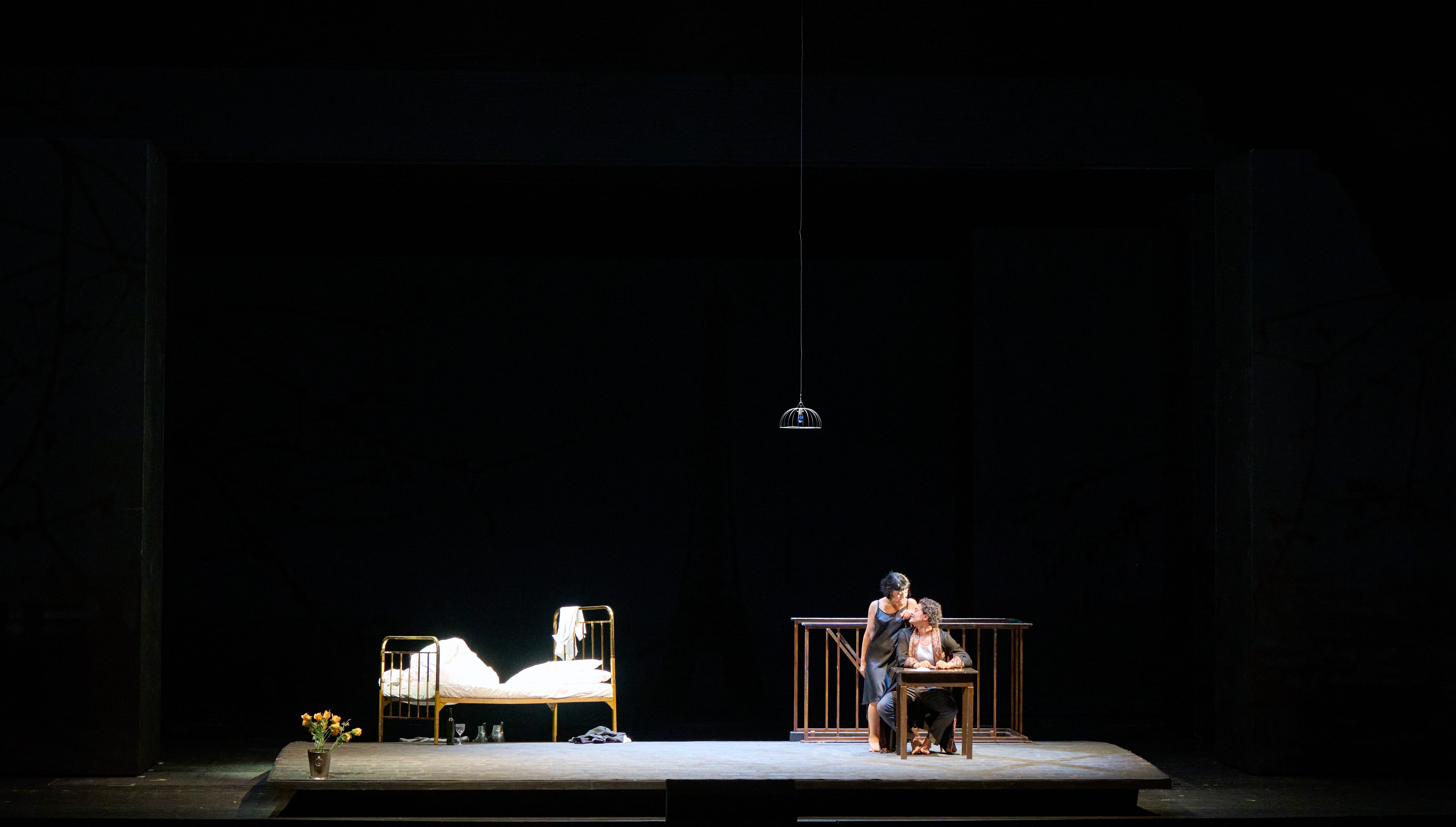 Zwei Personen sitzen eng umschlungen an einem Tisch auf einer spärlich beleuchteten Bühne; links steht ein leeres Bett unter einer Hängelampe.