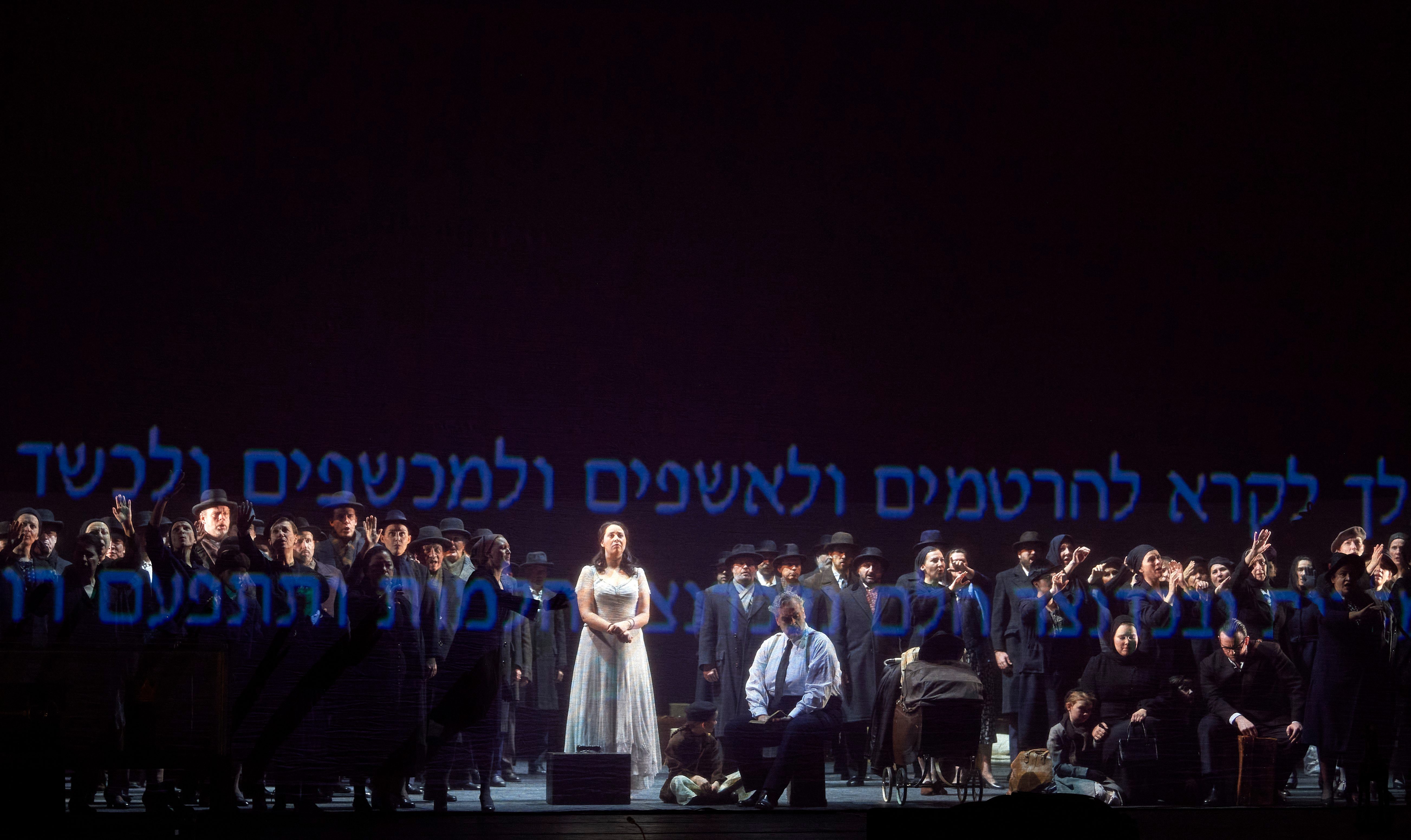 Eine Frau in hellem Kleid steht im Zentrum einer Gruppe dunkel gekleideter Menschen auf einer Bühne mit hebräischer Schriftprojektion.