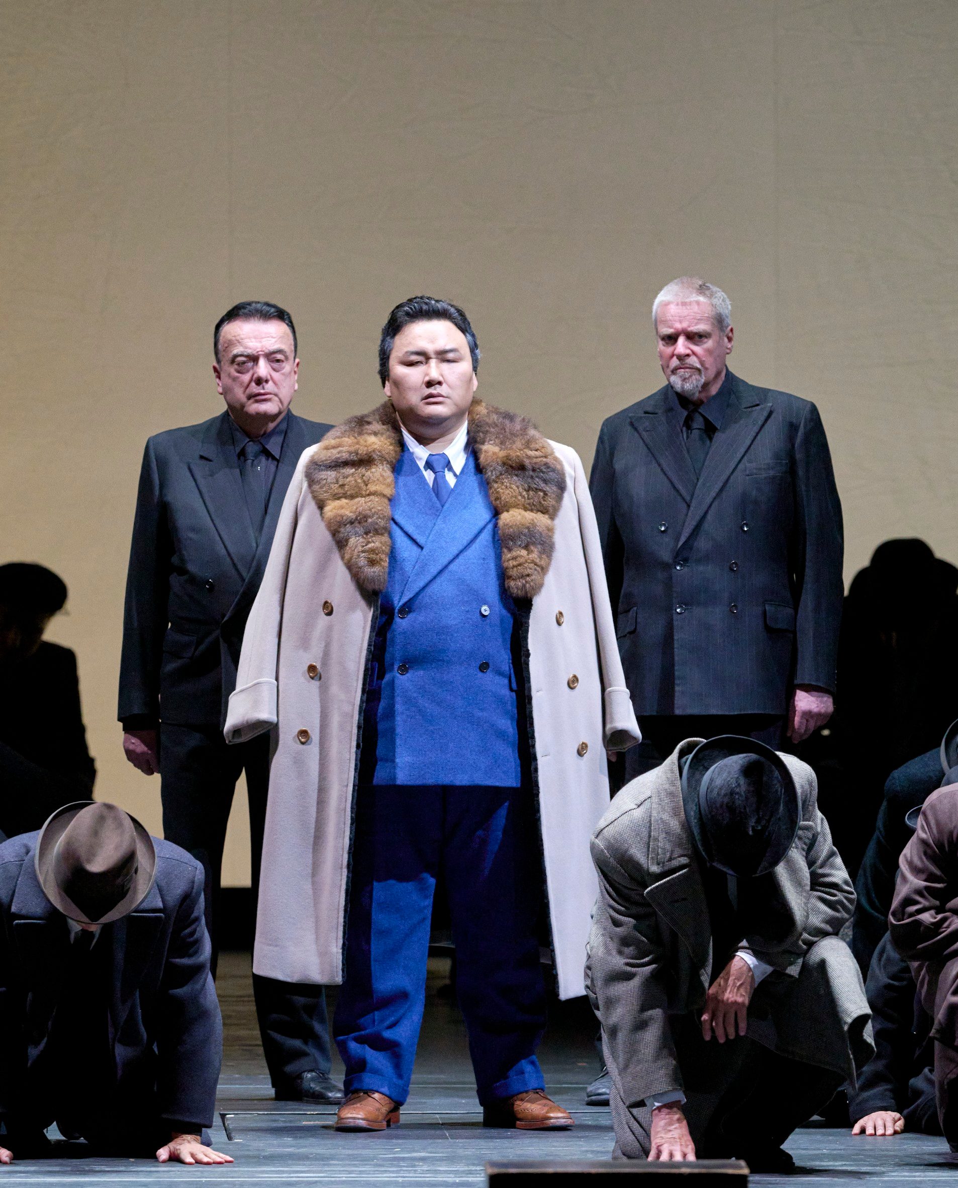 Ein Mann in königsblauem Anzug und Pelzmantel steht stolz zwischen zwei Männern in Schwarz, während andere sich vor ihm verbeugen.