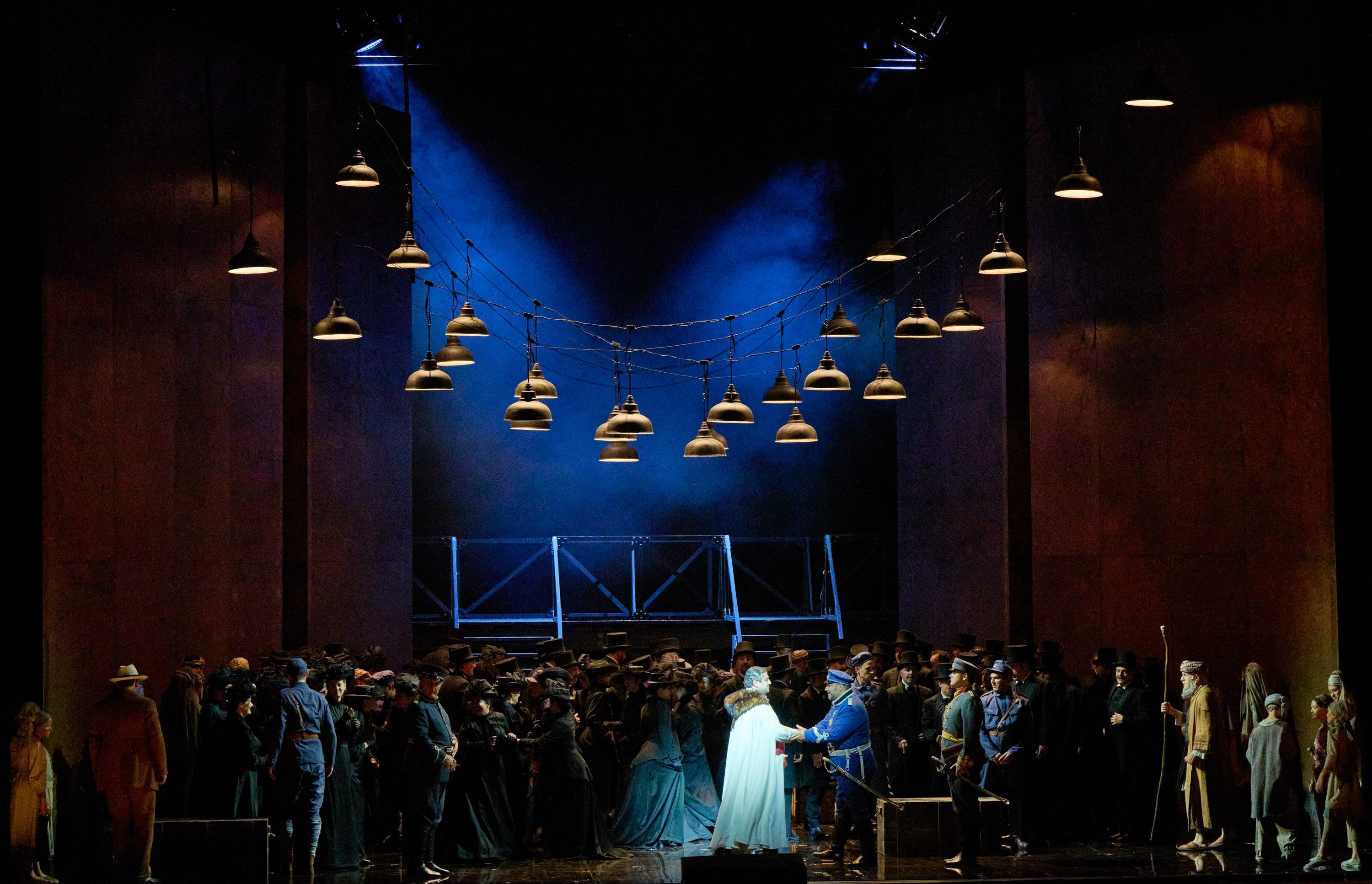 Eine Gruppe Menschen steht auf der Bühne. Über ihnen hängen viele Lampen. Im Zentrum stehen ein Mann in einem langen weißen Gewand und ein Mann in einer bauen Uniform.