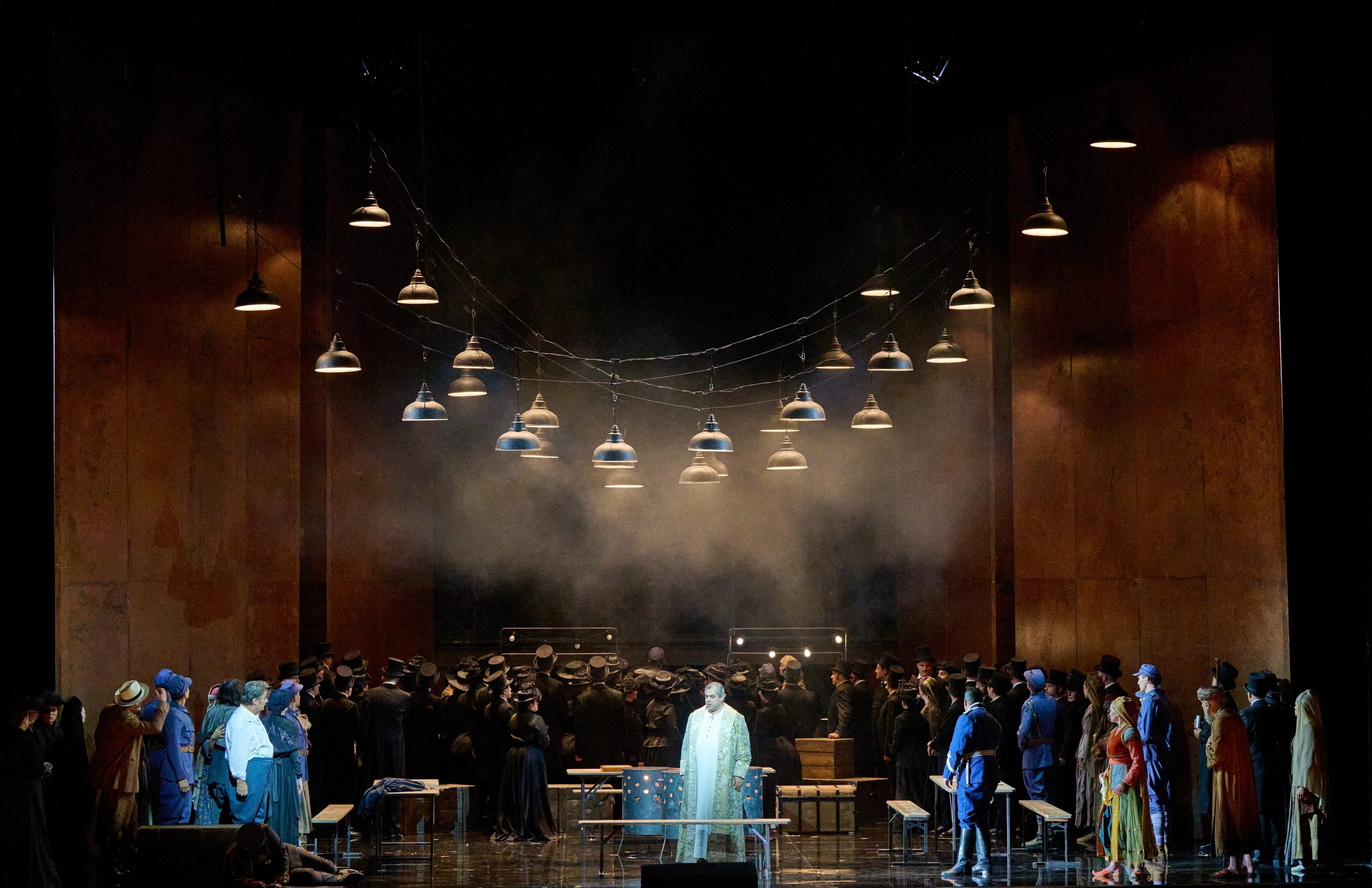 Eine Gruppe Menschen steht auf der Bühne. Über ihnen hängen viele Lampen. Im Zentrum steht ein Mann in einem langen weißen Gewand.