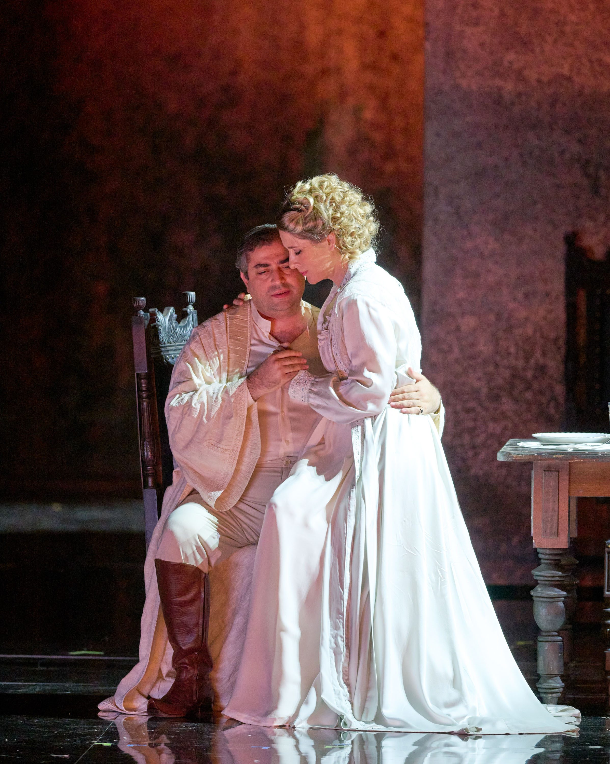 Ein Mann in einem weißen Gewand sitzt auf einem Stuhl. Auf seinem Schoss sitzt eine Frau in einem weißen Kleid.