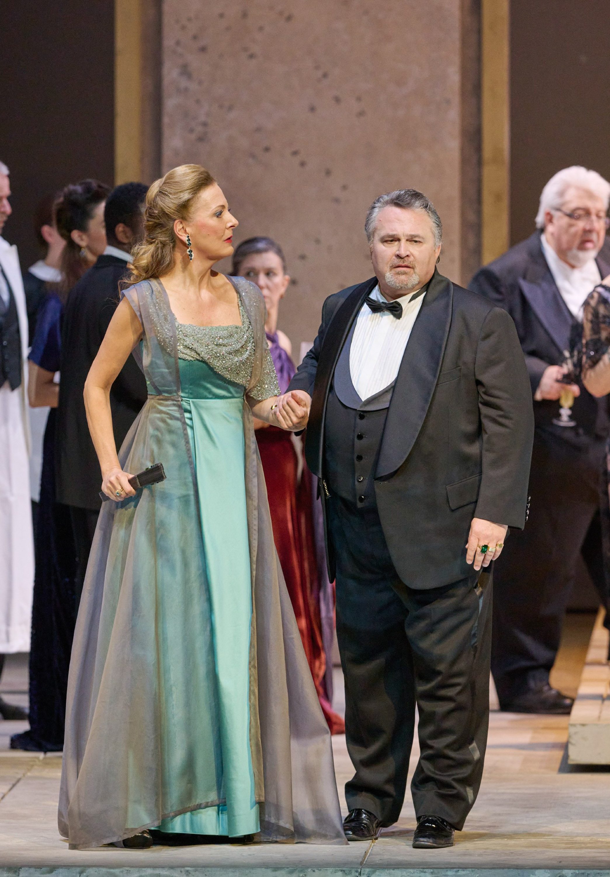 Eine elegant gekleidete Frau hält einen Mann im Smoking am Arm, während sie sich inmitten einer festlich gekleideten Gesellschaft befinden.