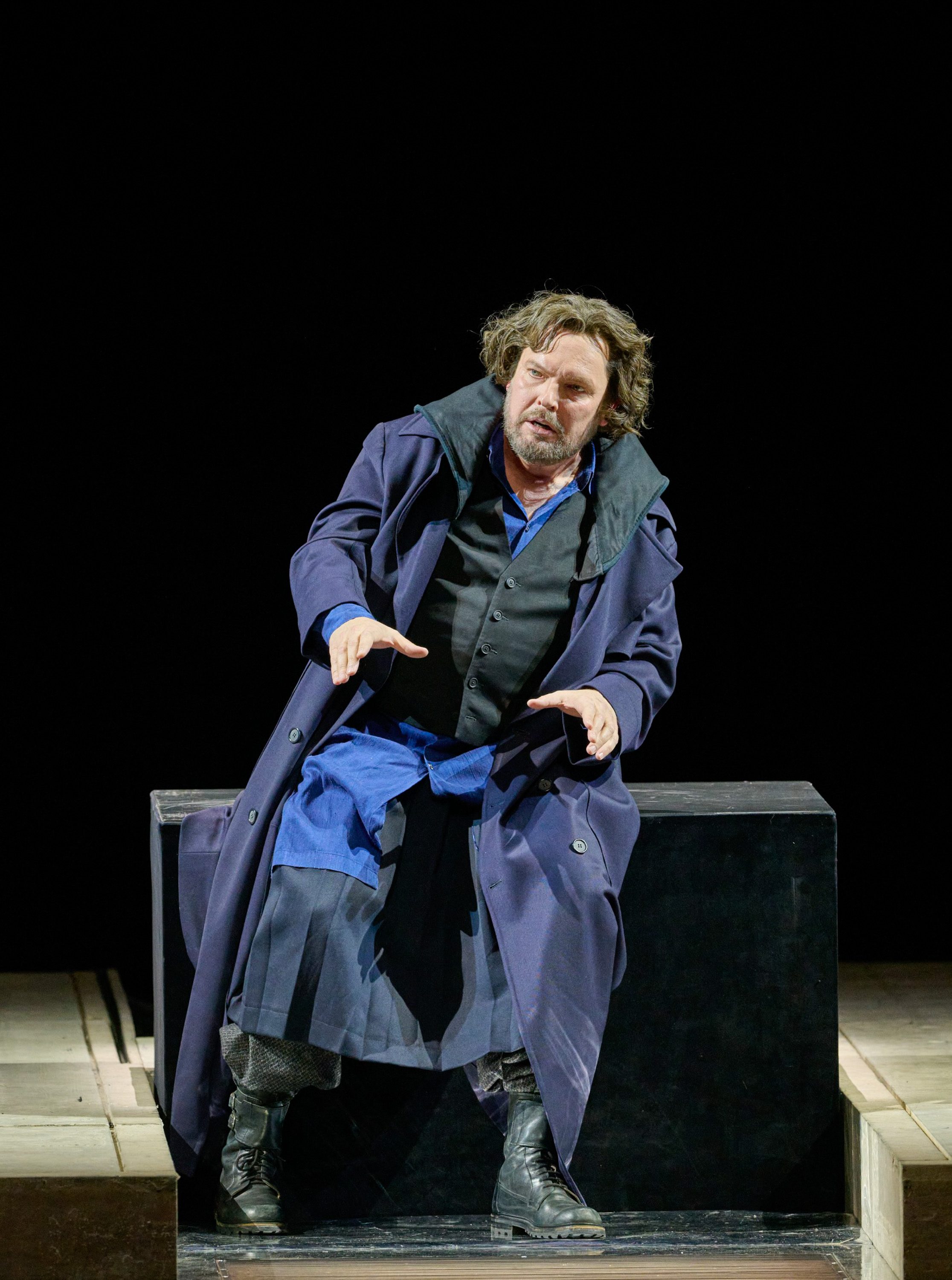 Ein Mann mit lockigem Haar sitzt in dunklem Mantel auf einer schwarzen Kiste und gestikuliert eindringlich vor schwarzem Hintergrund.