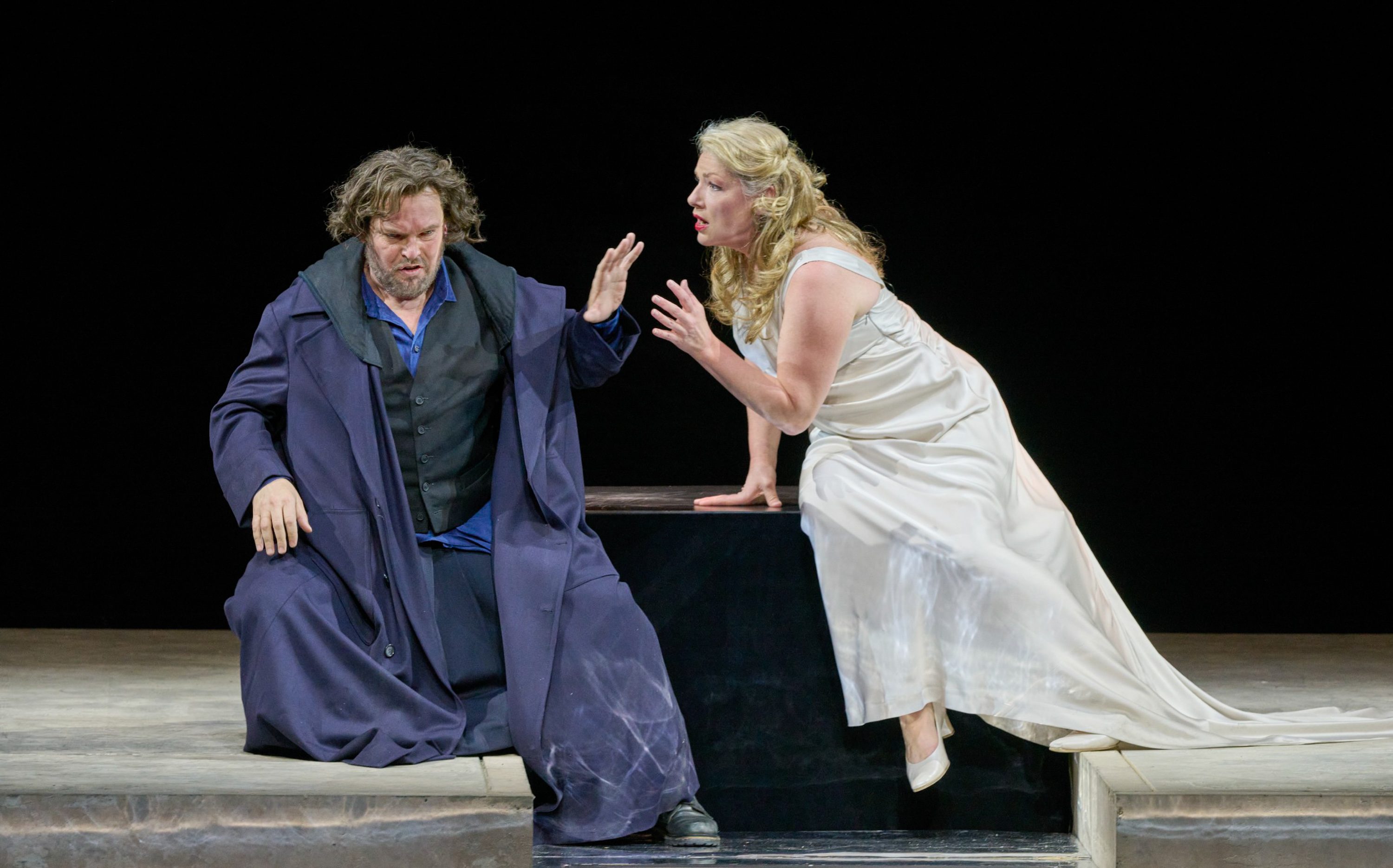 Ein Mann in dunklem Mantel wendet sich ab, während eine Frau im weißen Kleid aufgebracht auf ihn einredet.