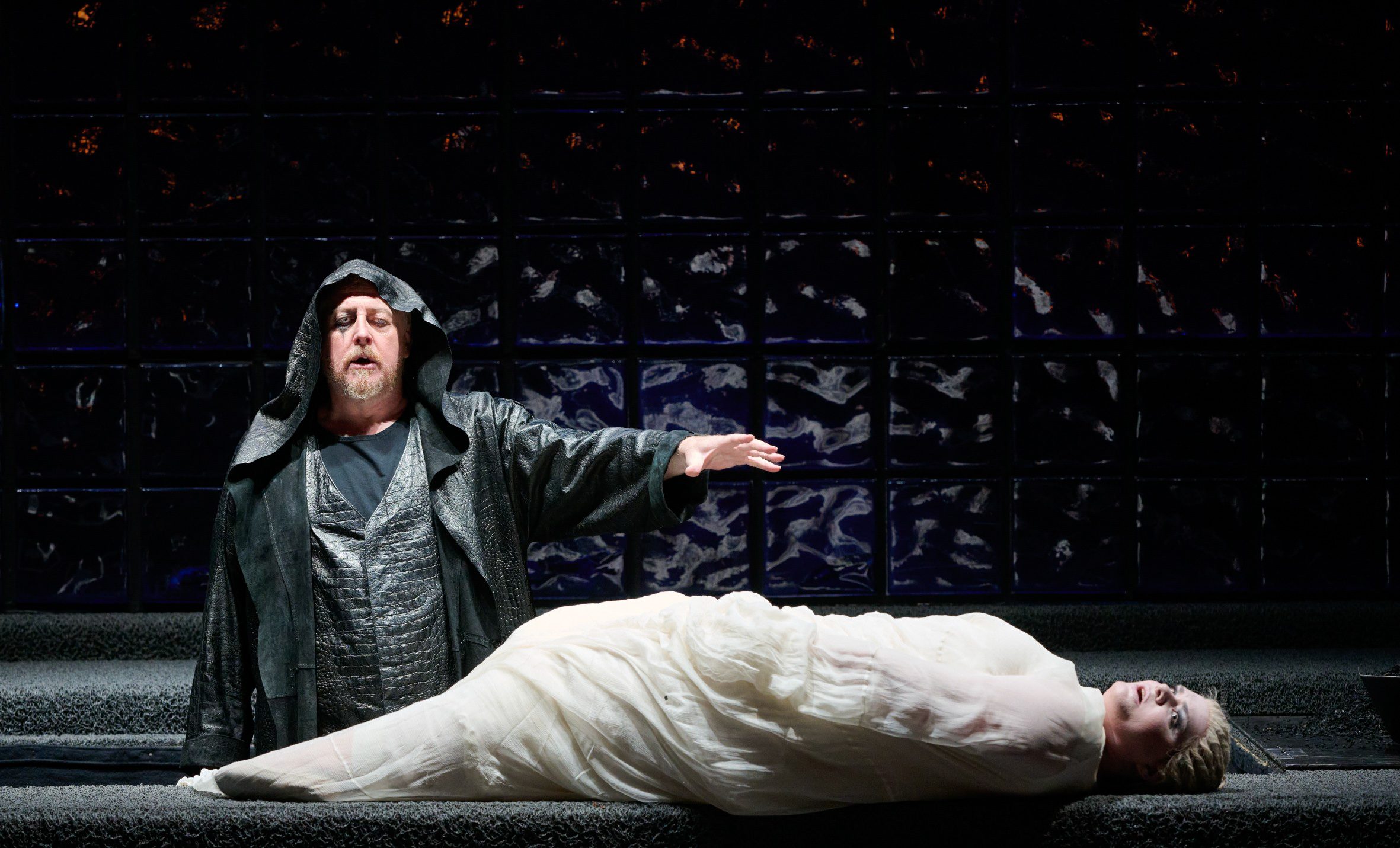 Ein Mann in schwarzem Mantel steht vor einer eingehüllten Person auf einer Liege.