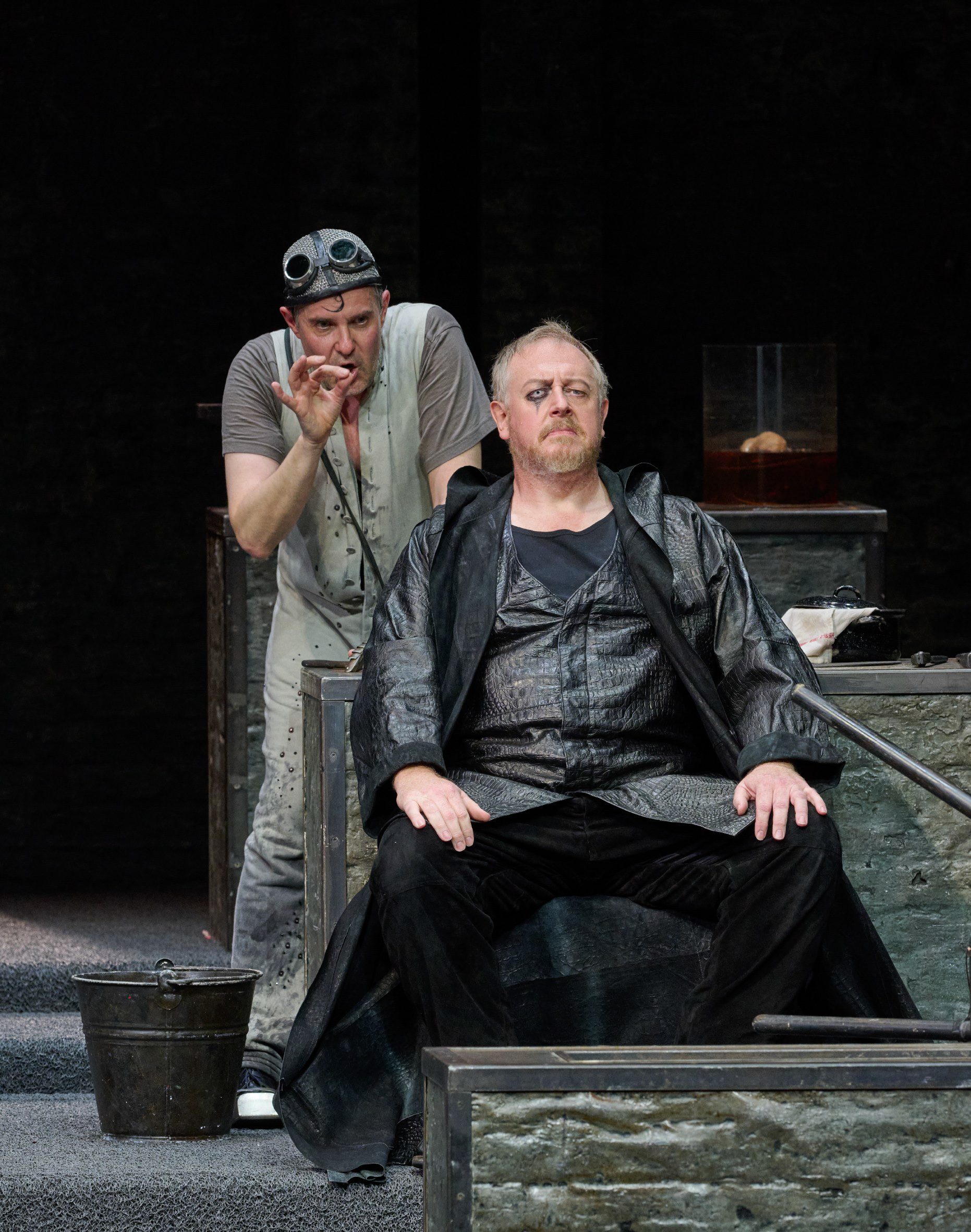 Zwei Männer vor dunklem Hintergrund; der eine sitzt, der andere steht dahinter und beide blicken nach vorne.