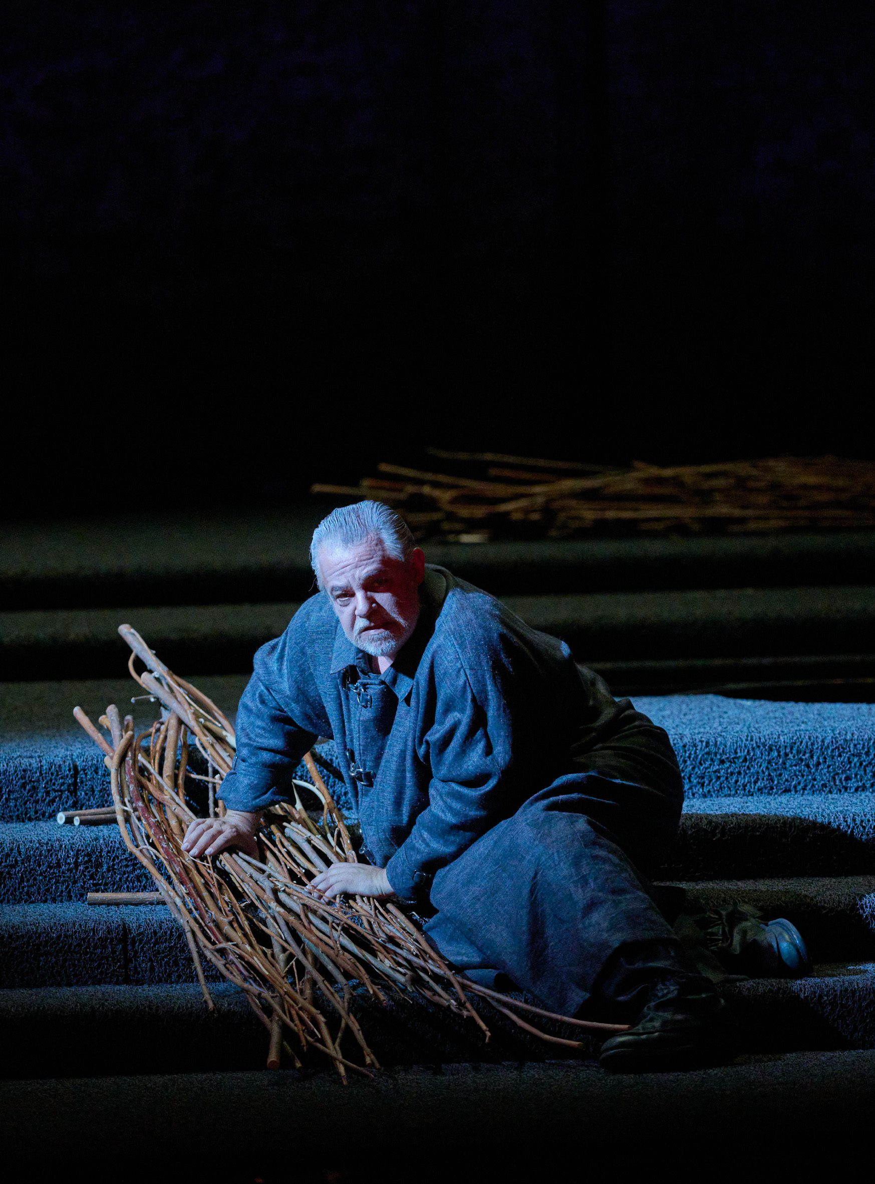 Ein Mann in grauer Kleidung sitzt auf einer Treppe, umgeben von Zweigen.