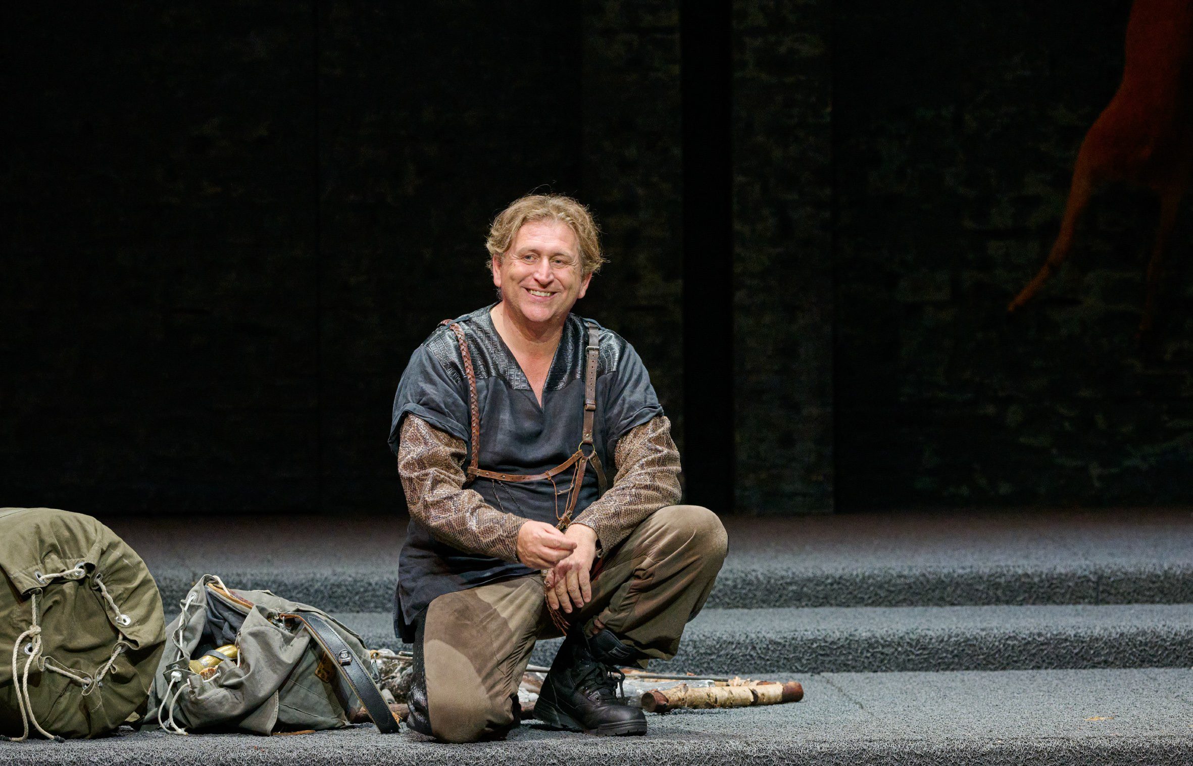 Ein Mann in dunkler Kleidung kniet auf einer leeren Bühne und blickt nach vorne.