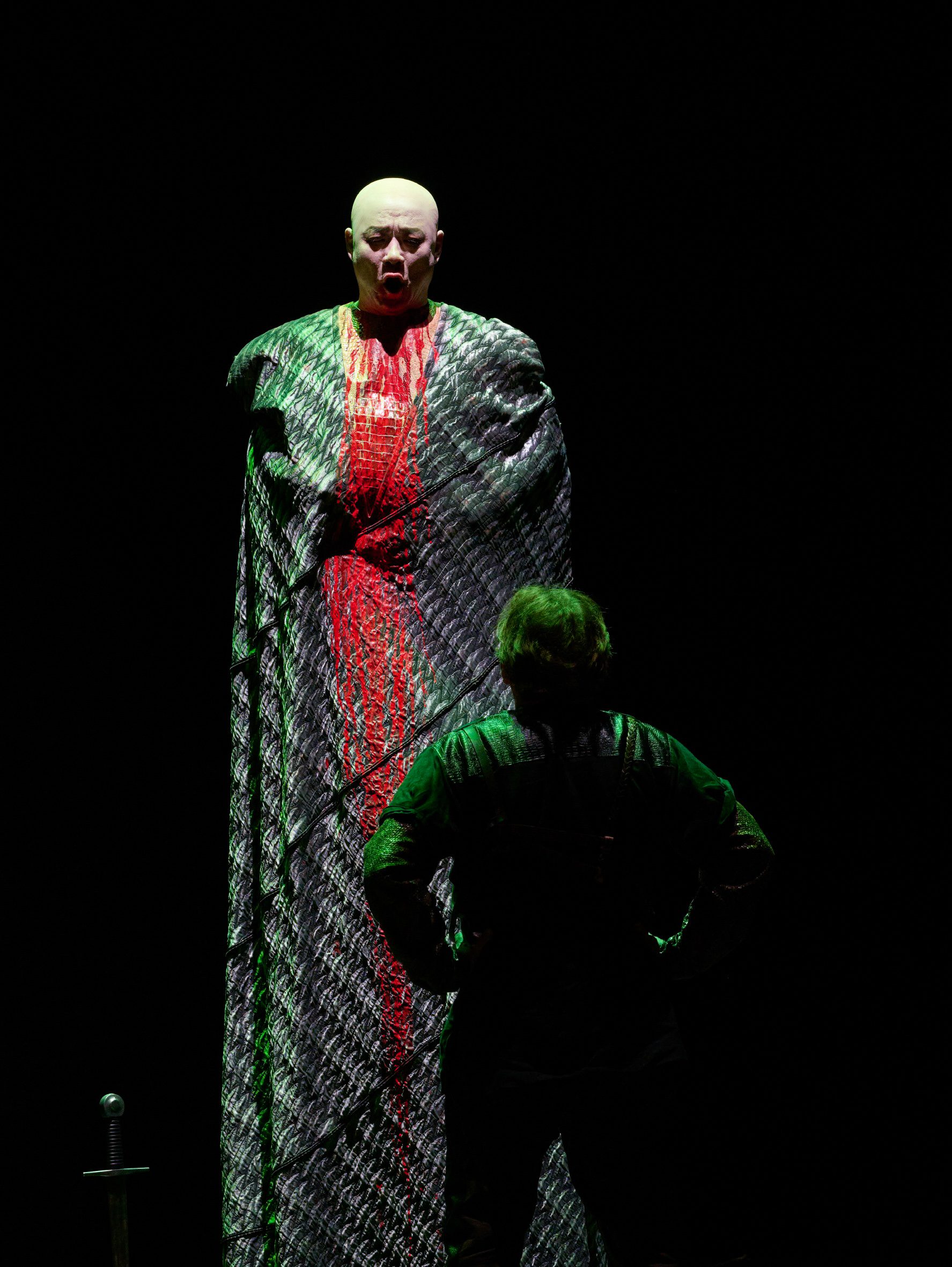 Eine große Gestalt im langen Mantel spricht zu einem vor ihm stehenden Mann.