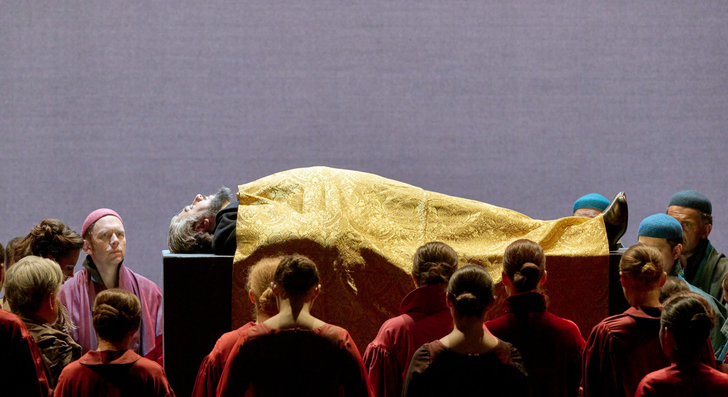 Eine Gruppe Menschen steht um eine erhöhte Plattform, auf der ein Mann mit goldenem Umhang und zurückgelehntem Kopf liegt.