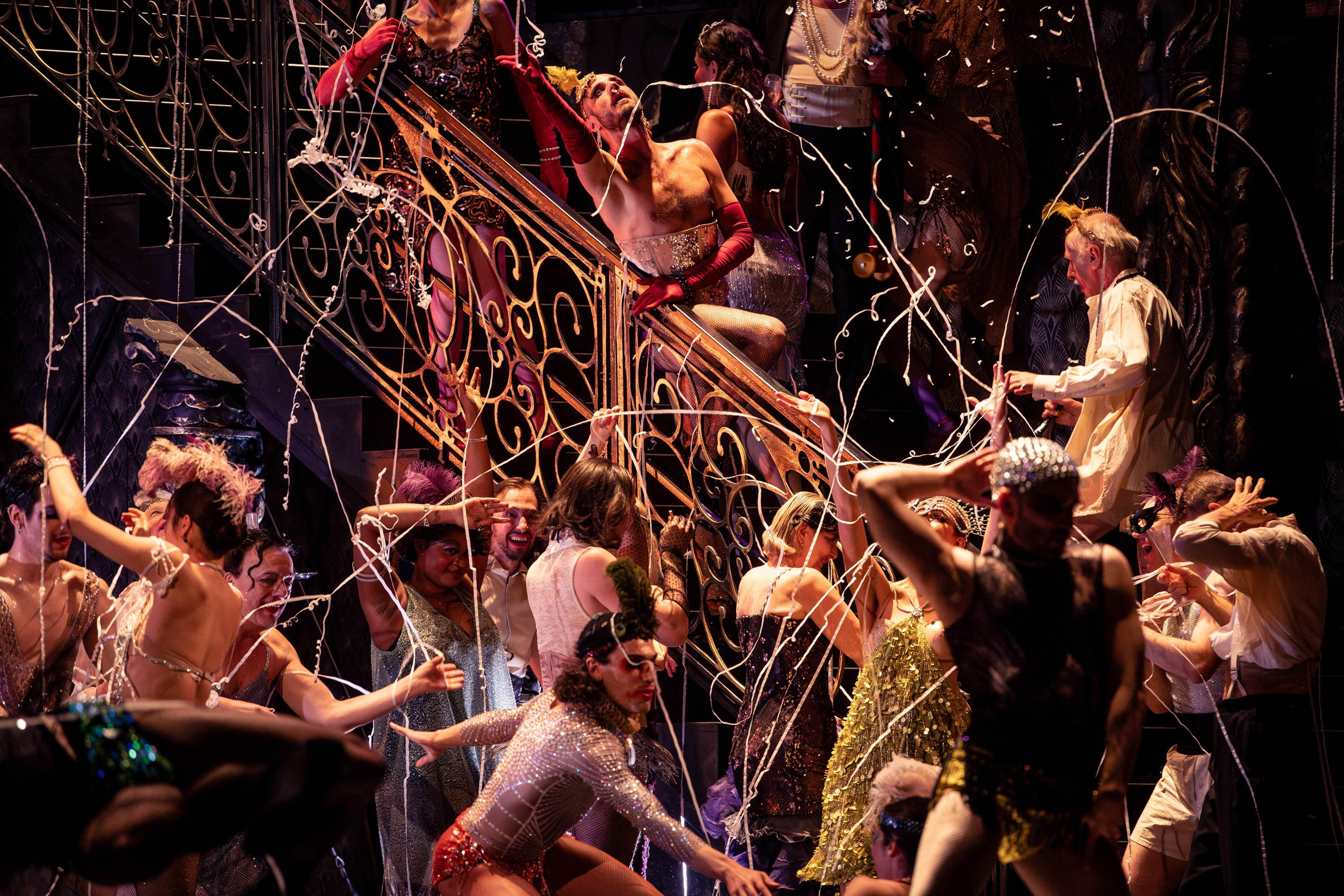 Viele Männer in Glitzerkostümen stehen auf und neben einer Treppe. Es Fliegen Luftschlangen umher.