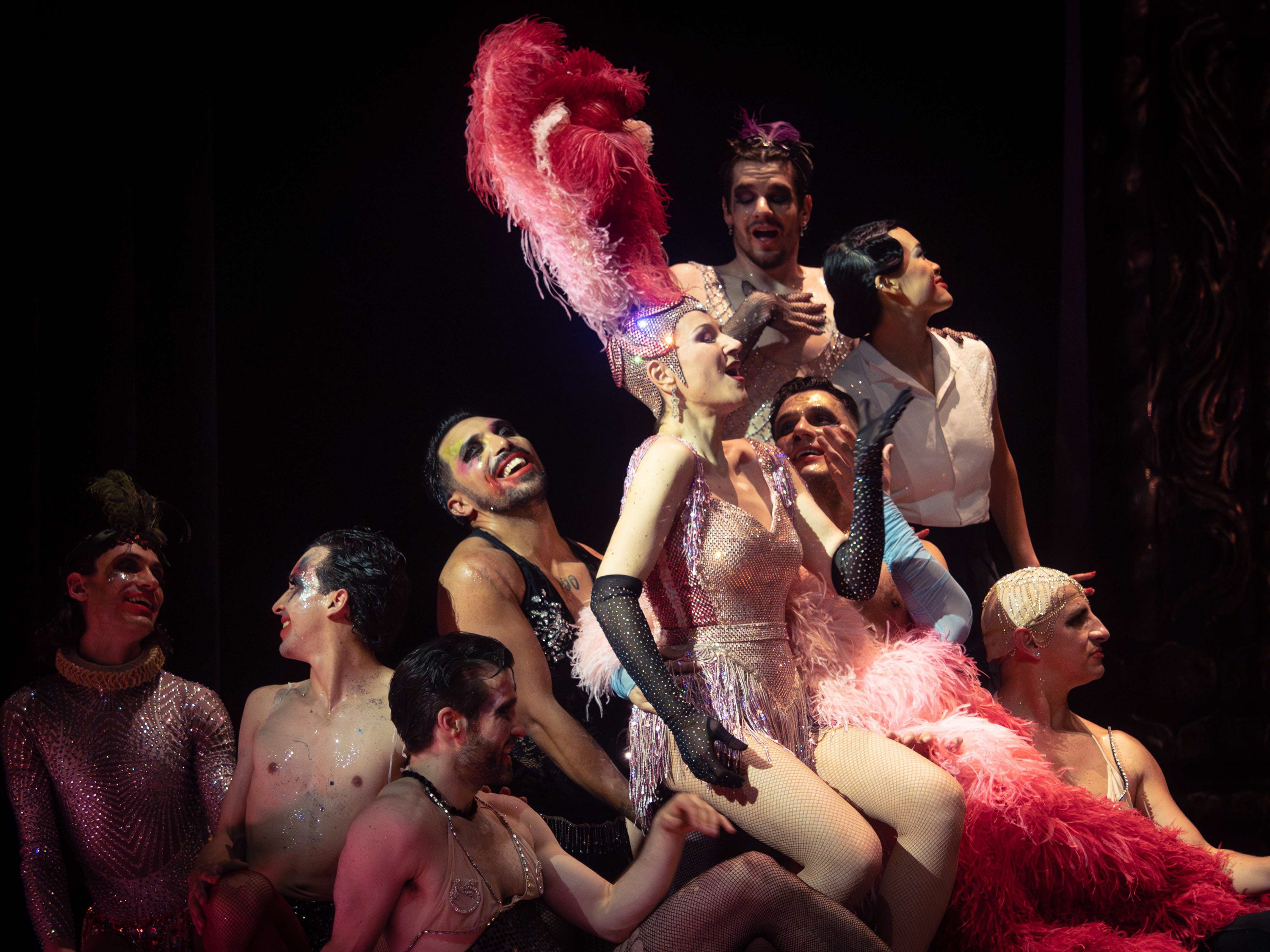 Eine Frau in einem pinken Glitzerkostüm und einer Federkrone auf dem Kopf sitzt auf einem Haufen Männern