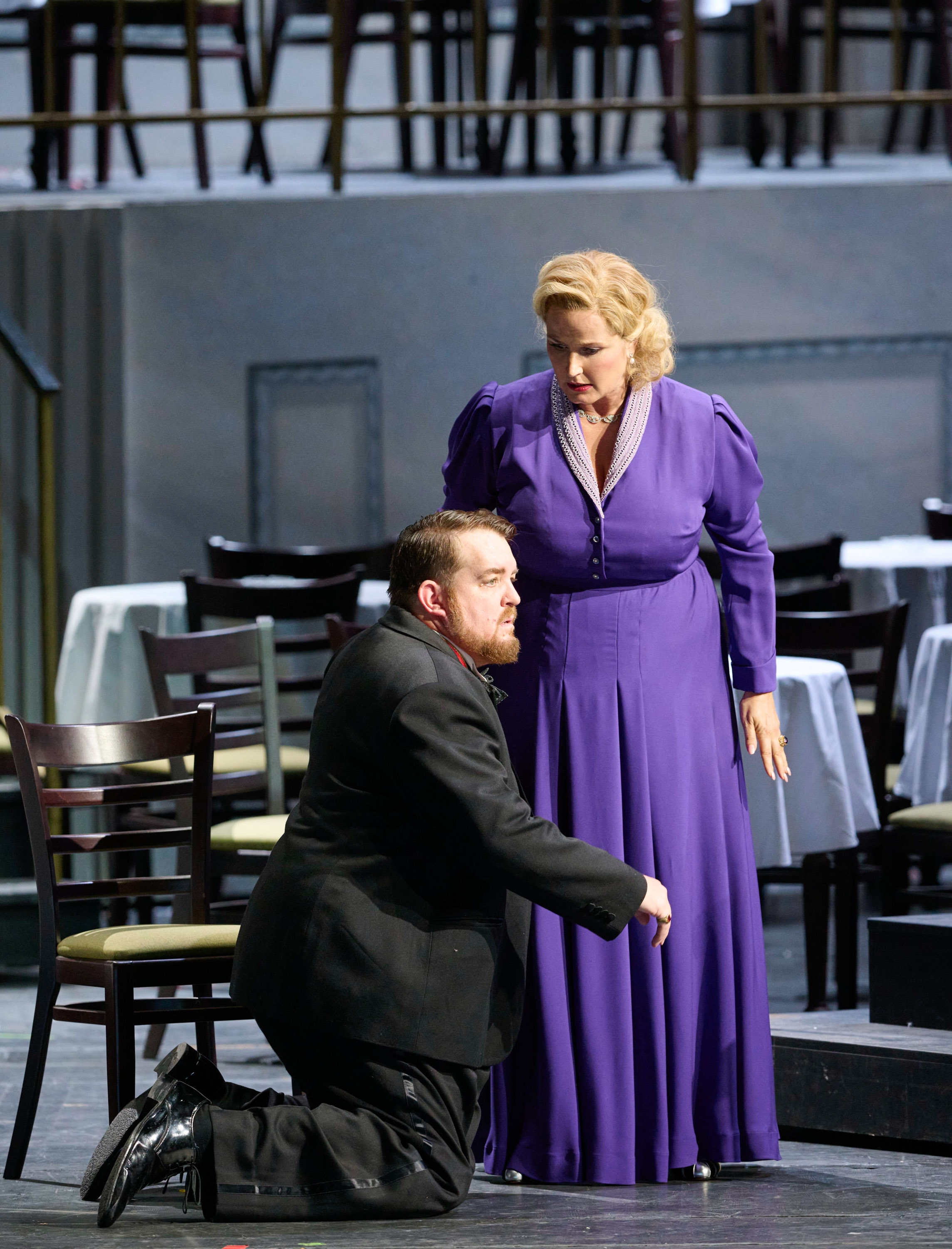 Eine blonde Frau in einem langen violetten Kleid steht inmitten von Tischen und Stühlen. Zu ihren Füßen Kniet ein Mann, der einen Smoking trägt.