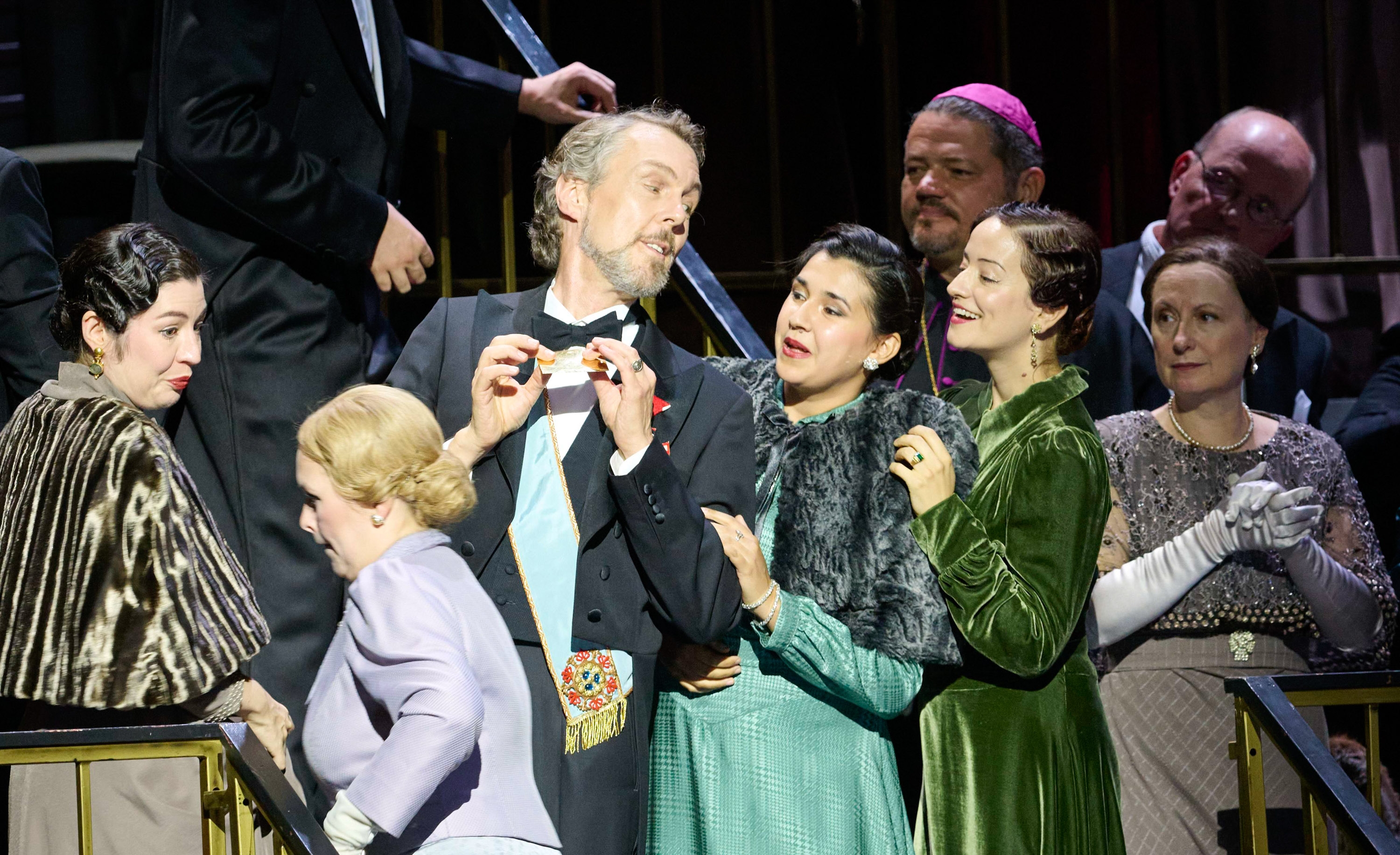 Man sieht einen Mann in einem Smoking und zwei Frauen. Die eine trägt ein türkises Kleid.